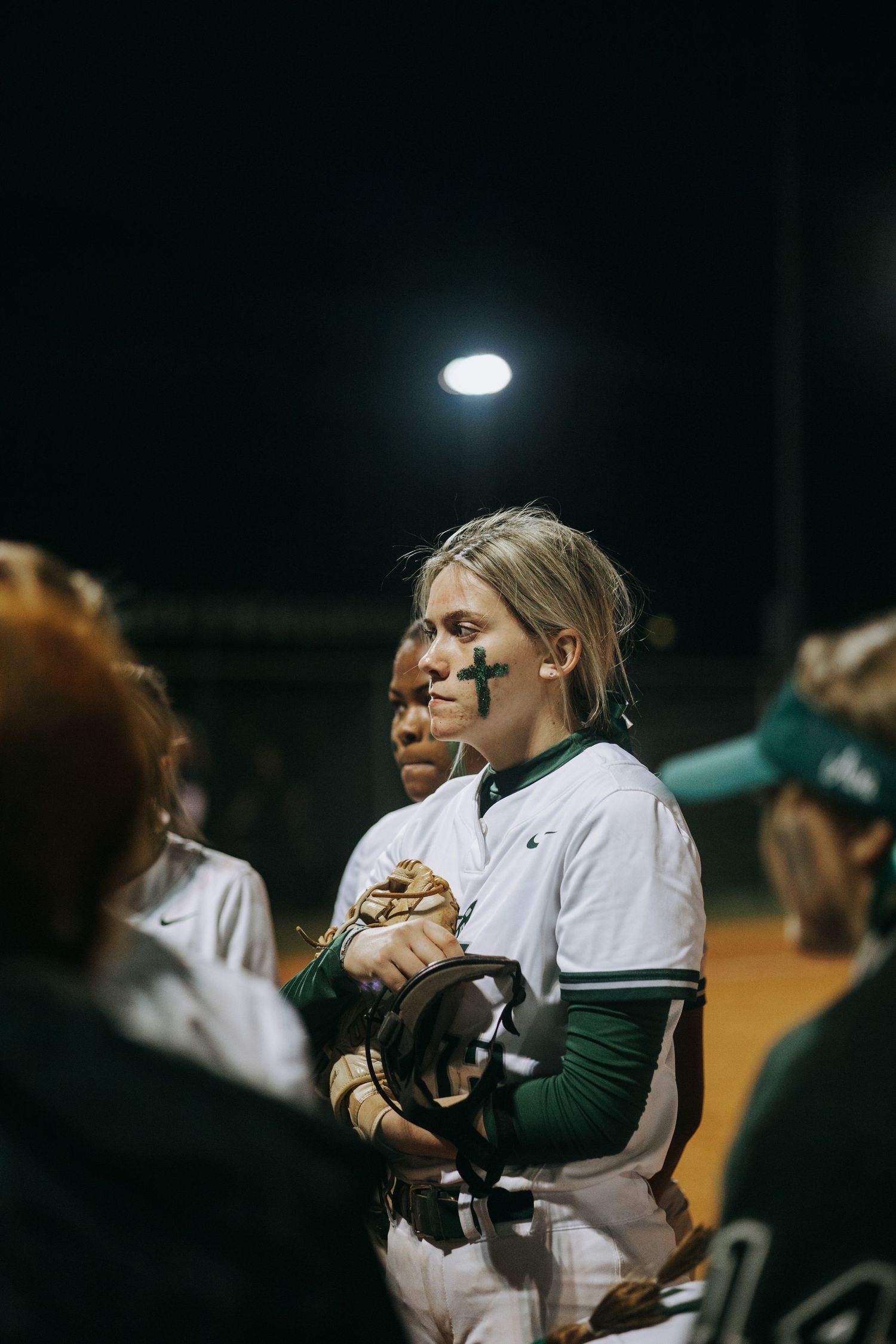 Softball player in white and green uniform smiles at night game under stadium lights.