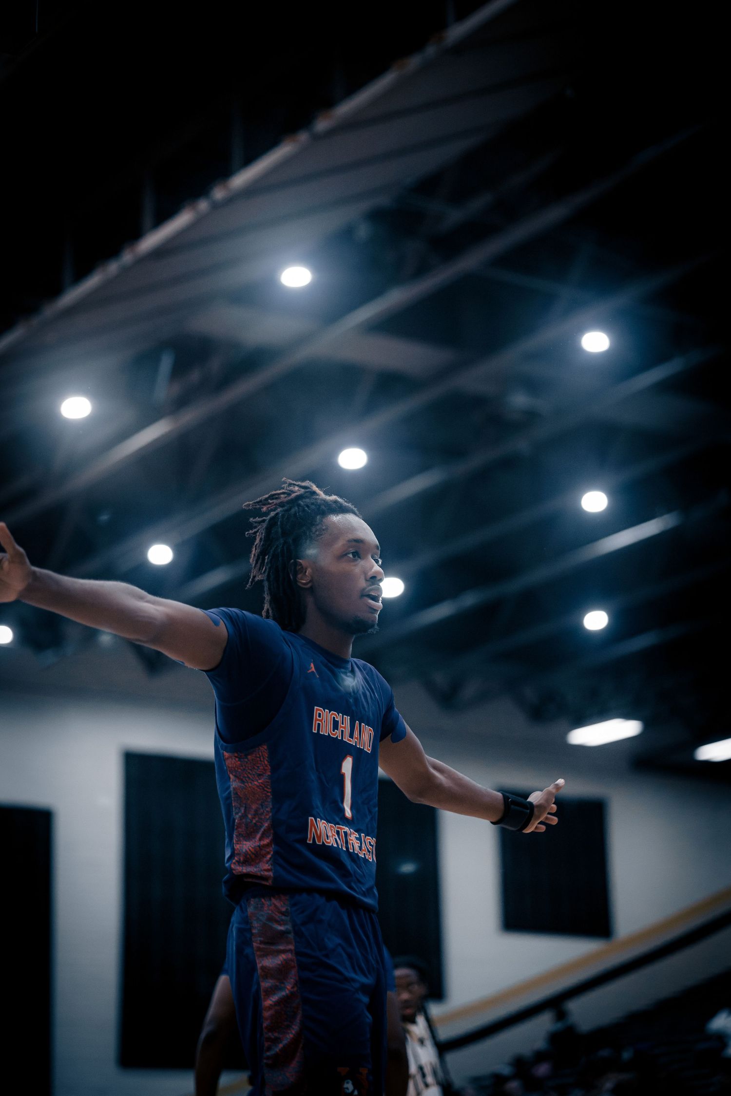 Basketball player in blue jersey gestures with arms outstretched during indoor game under bright lights.
