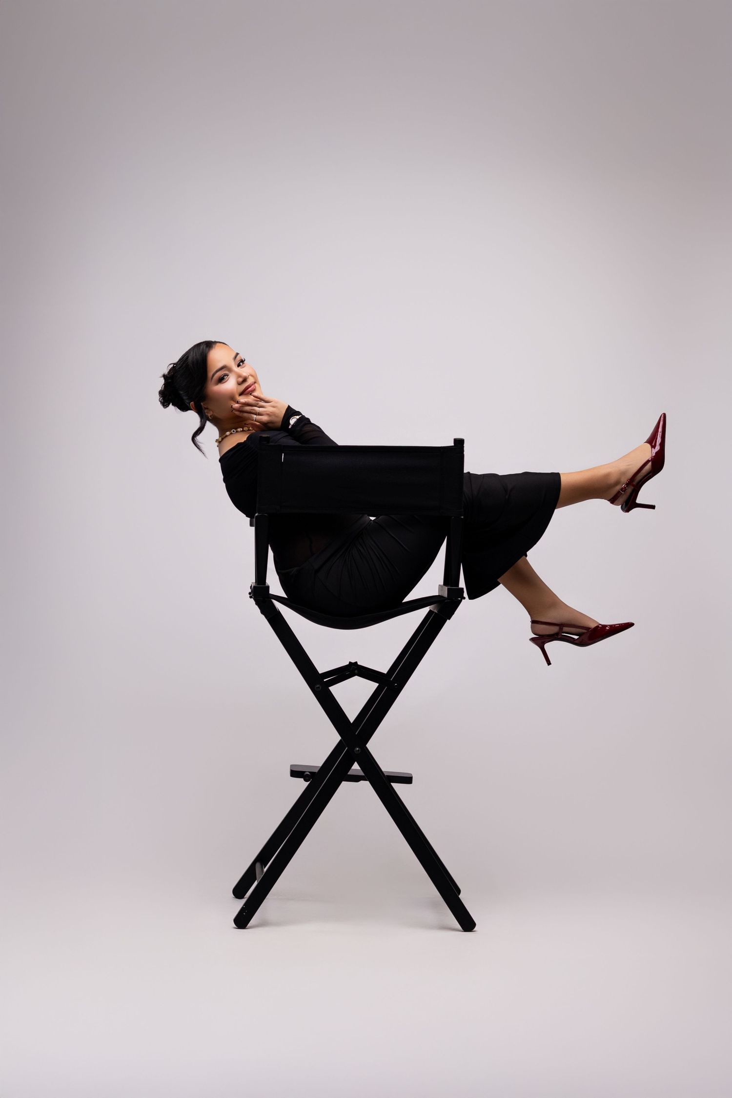 Stylish figure in black dress lounging elegantly on director's chair against light background.
