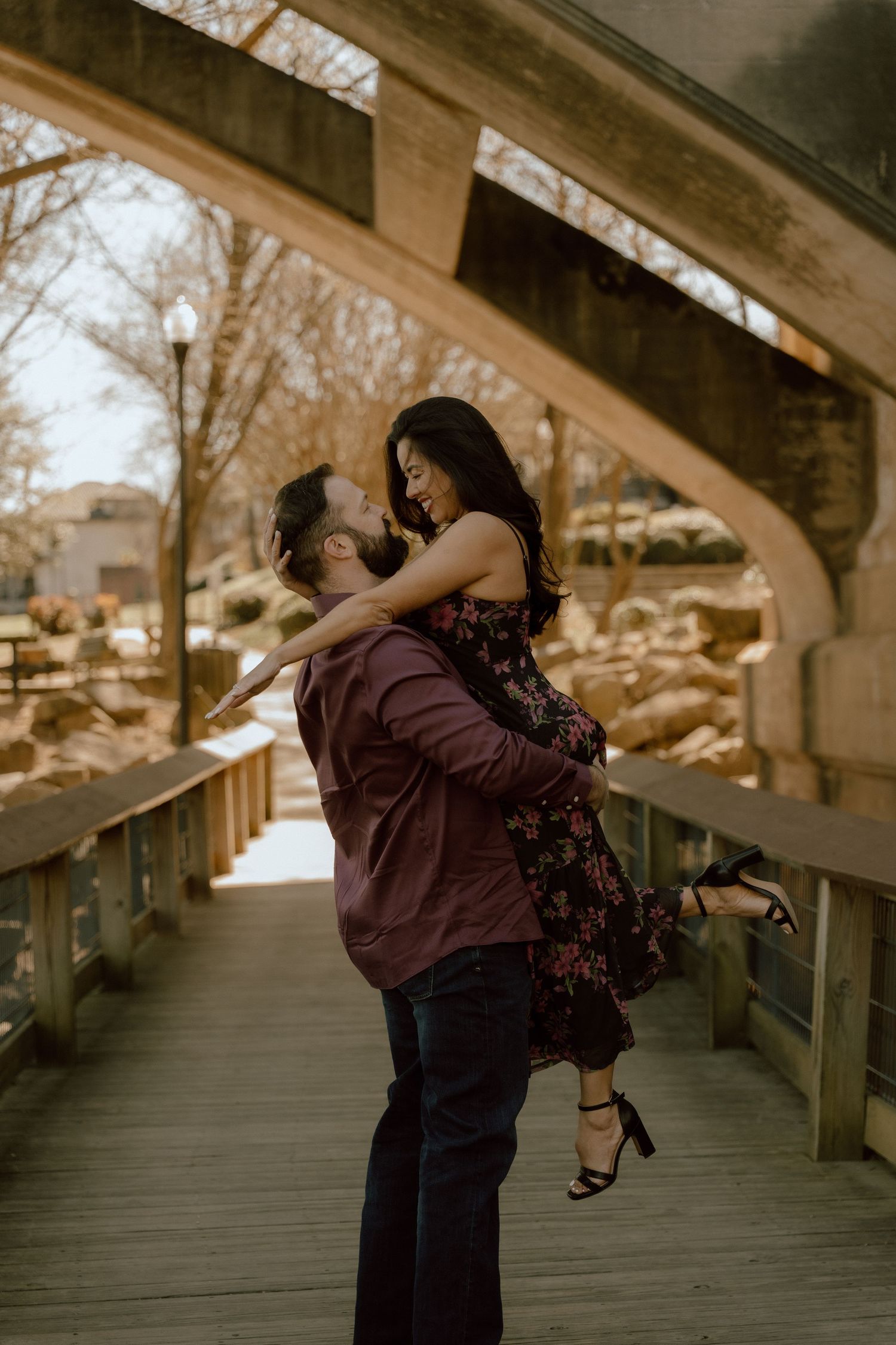 A man lifts a woman in a floral dress on a wooden bridge, sharing a romantic moment outdoors.