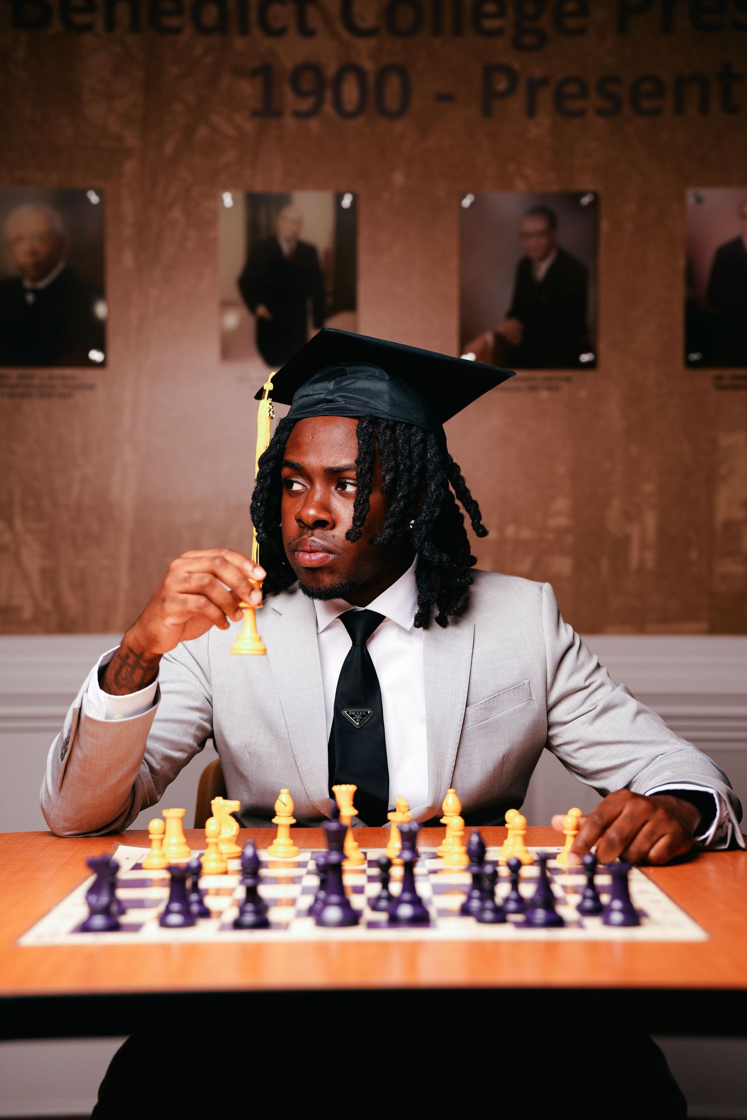 Graduate in cap and suit plays chess at Benedict College, holding a piece before portraits of presidents.