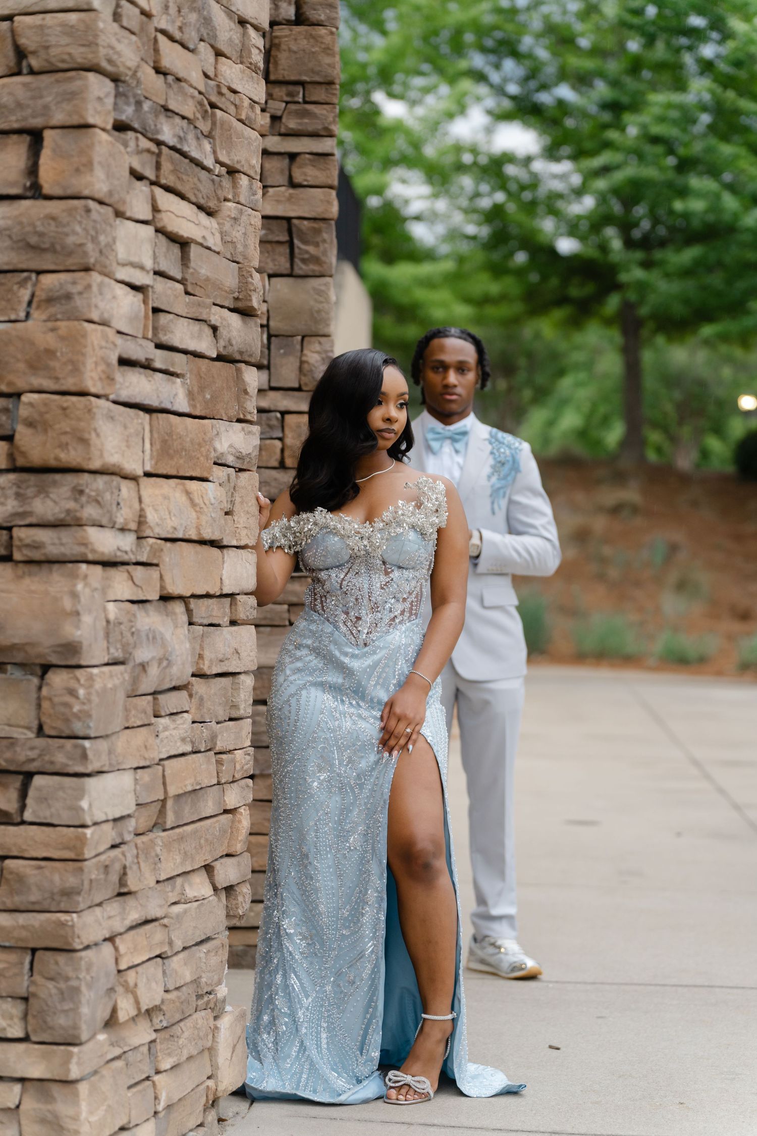 A couple poses elegantly in prom attire, she in a silver sequin gown and he in a white suit with blue bow tie.