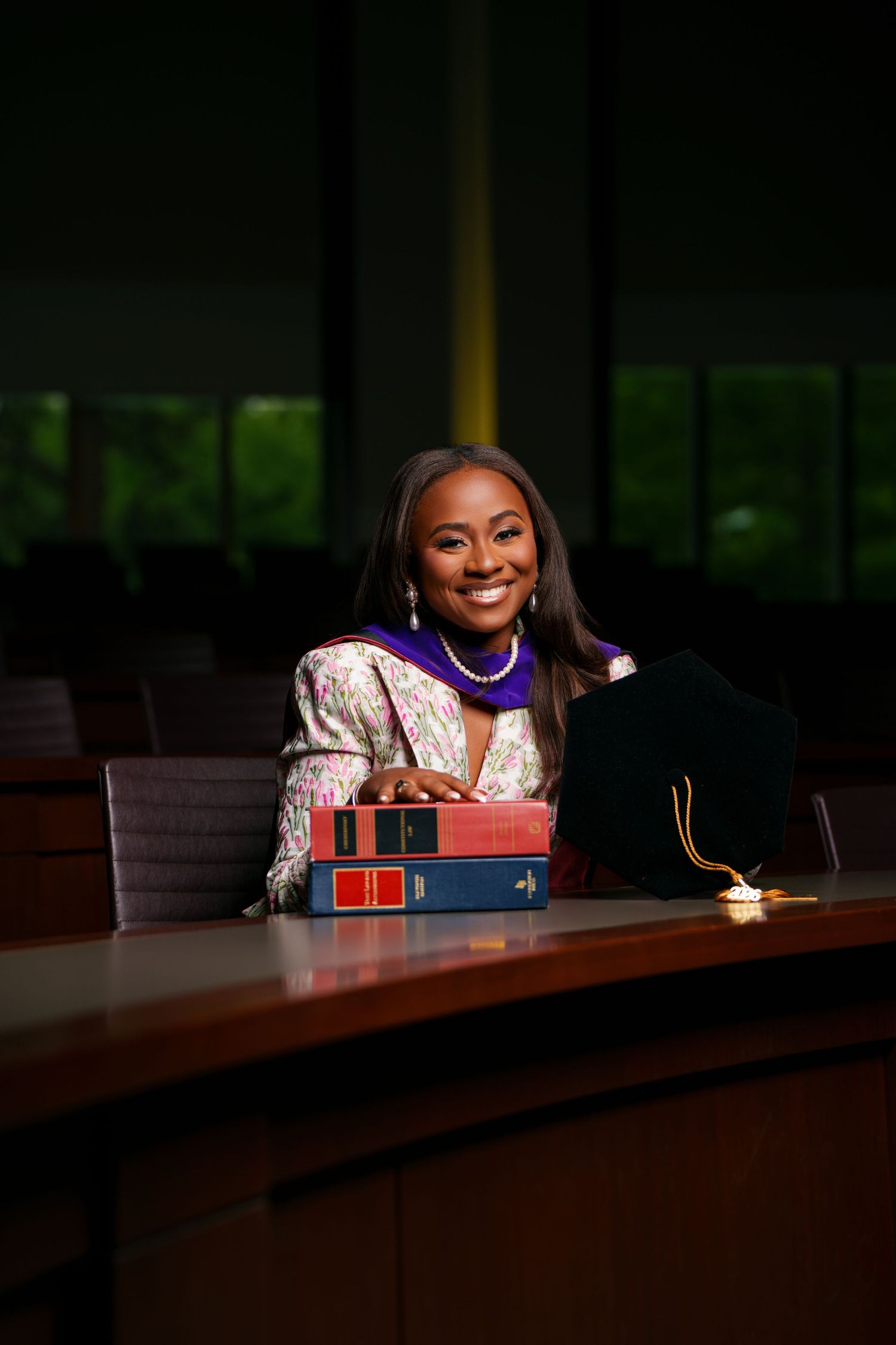 Smiling woman in floral blazer poses with law books and graduation cap at a courtroom desk.