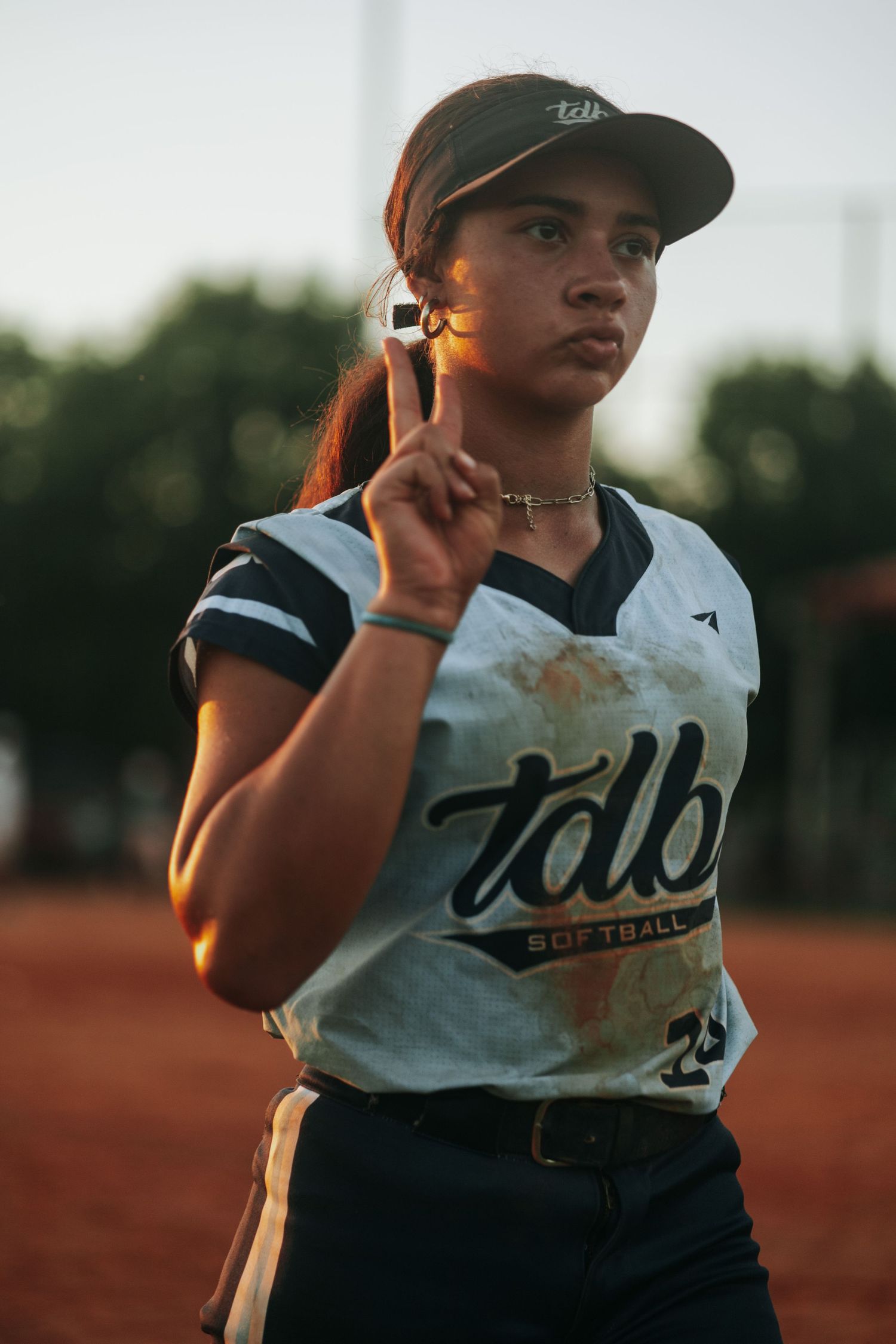 A softball player in a white uniform stands ready during golden hour.