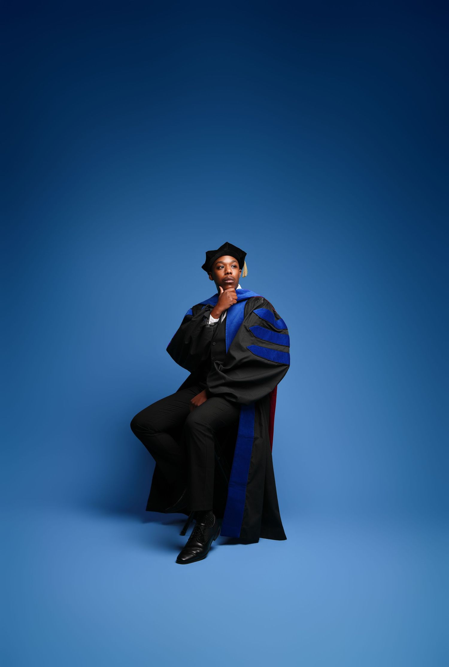 Graduate in doctoral regalia poses confidently in black and blue gown with cap against blue studio background.
