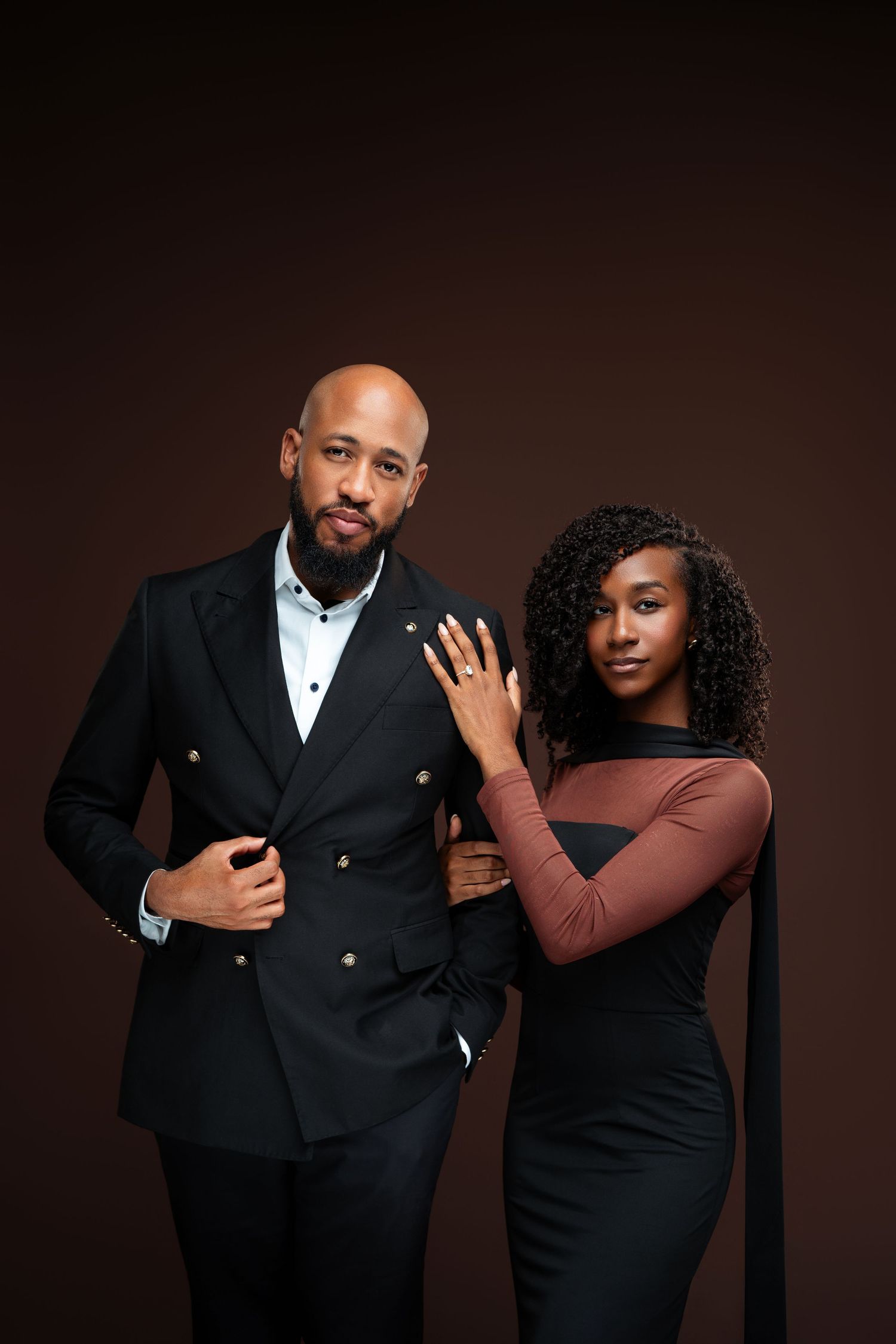 Elegant couple posing in studio, man in black double-breasted suit, woman in brown and black dress, dark background.