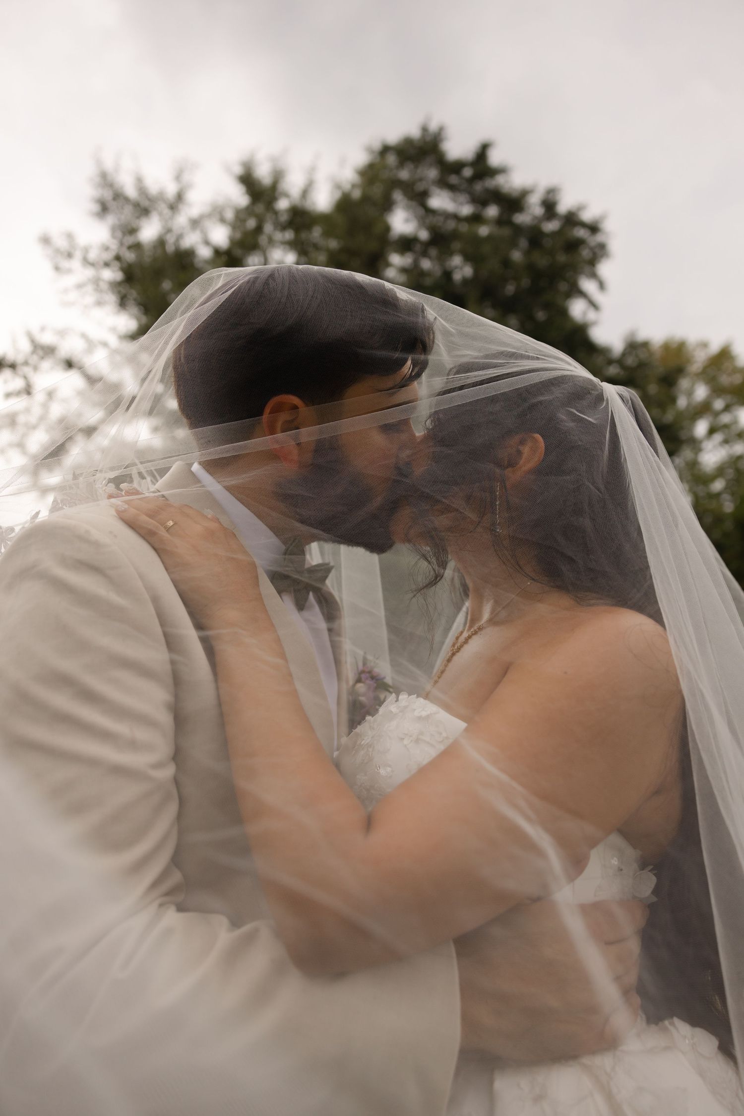 Bride and groom sharing a romantic kiss under a flowing white veil outdoors on their wedding day.