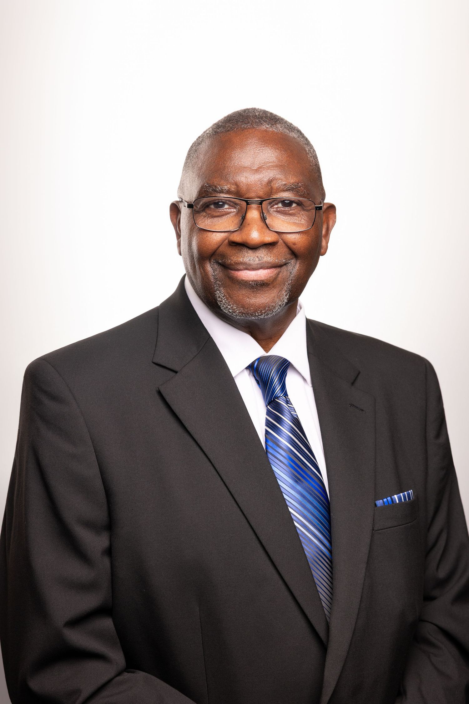 Professional headshot of a smiling senior man in a black suit, blue striped tie, and glasses against a white background.