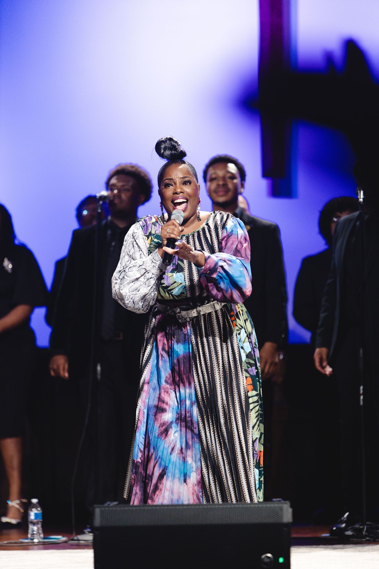 Performer in patterned dress sings energetically on stage with blue lighting.