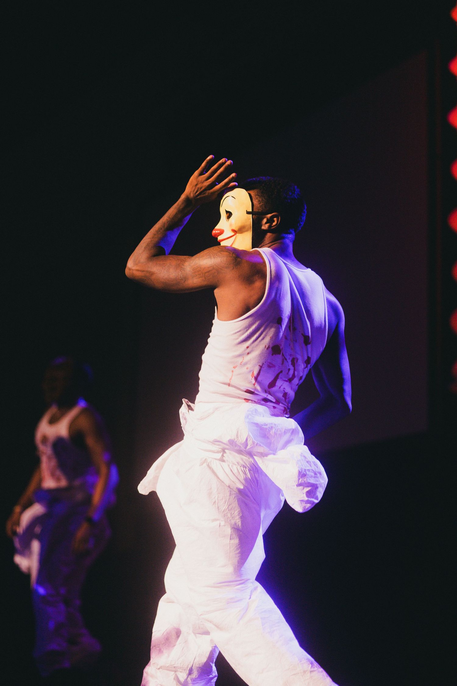 question perform in white costumes against a dark background illuminated by dramatic purple stage lighting.