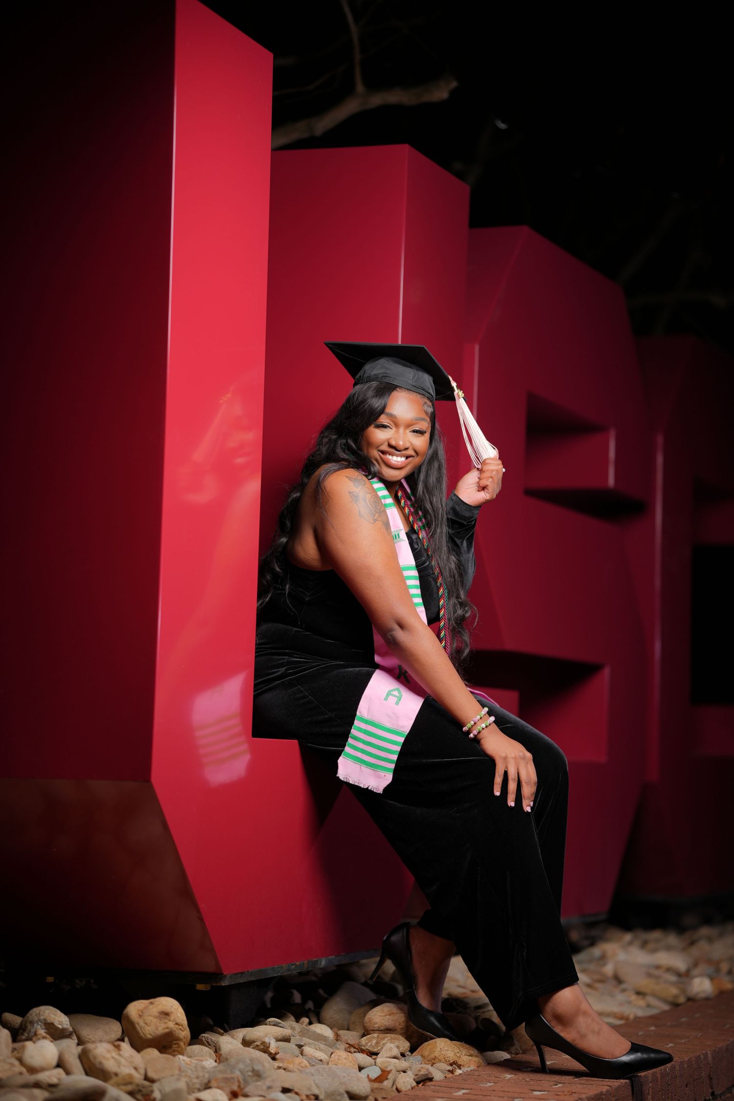 A woman in a black dress poses against a red wall background while sitting on a floor with rocks.