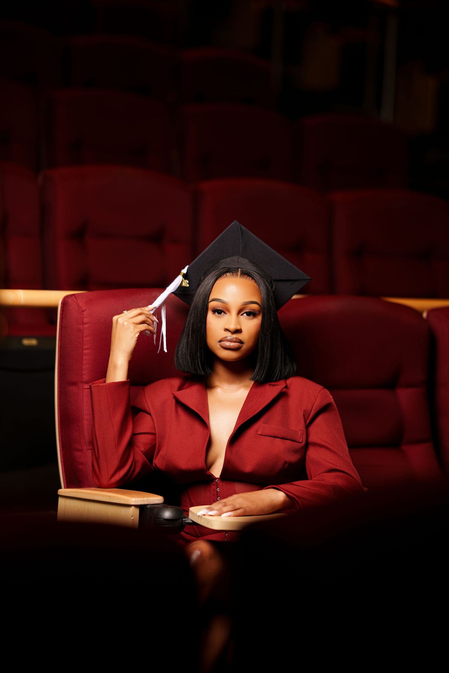 Graduate in red outfit wearing mortarboard cap seated in auditorium.