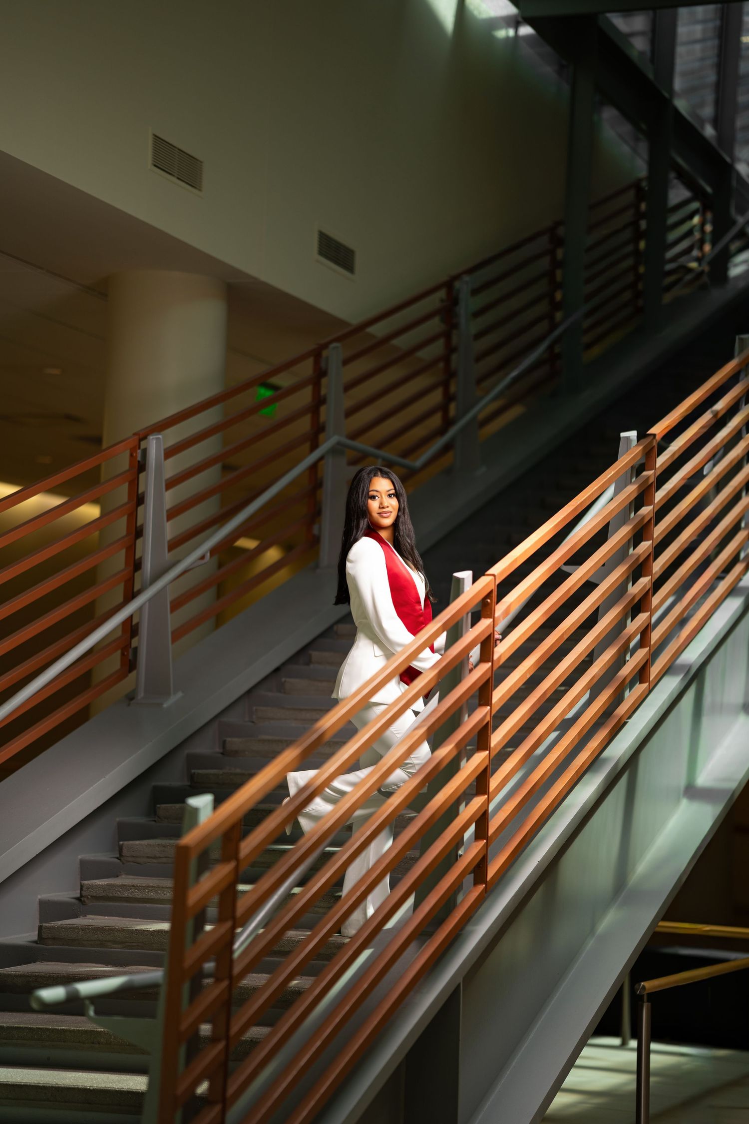 A young woman in a red and white outfit stands on modern stairs with wooden railings in a contemporary building.