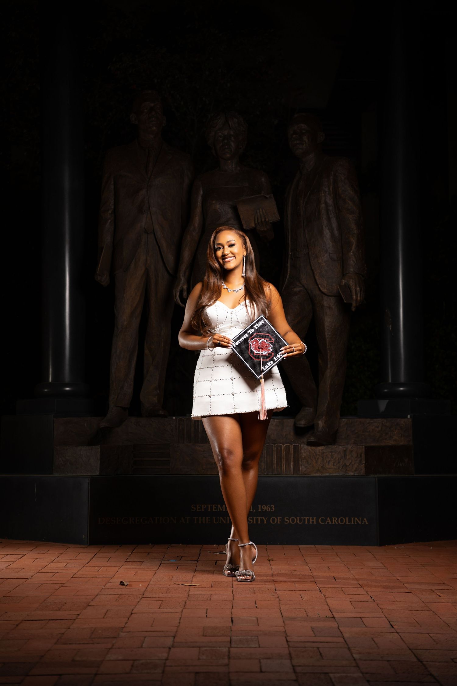 A smiling graduate in a white dress holds a University of South Carolina diploma in front of bronze statues at night.