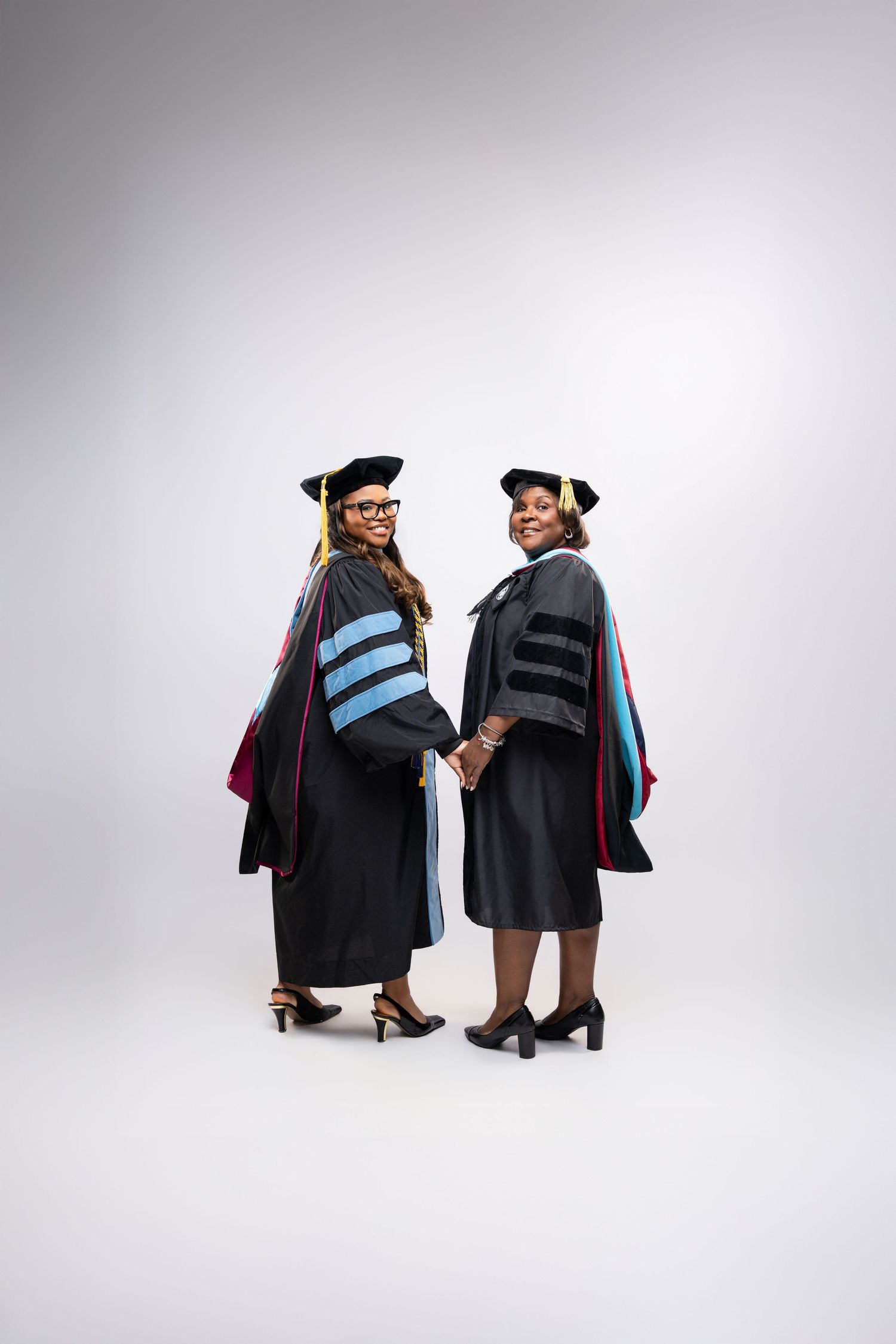 Two women in doctoral graduation gowns and caps holding hands, smiling over their shoulders in a studio portrait.