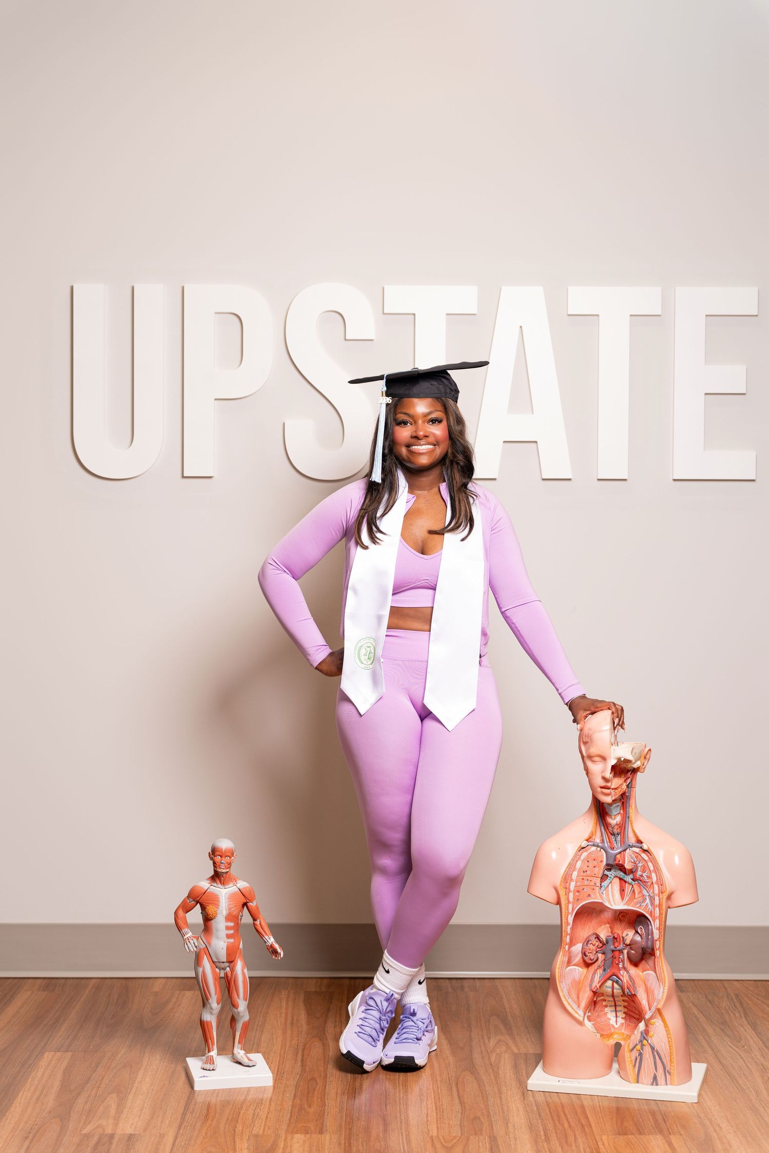 Smiling graduate in purple outfit and cap poses with anatomy models in front of UPSTATE sign on wall.