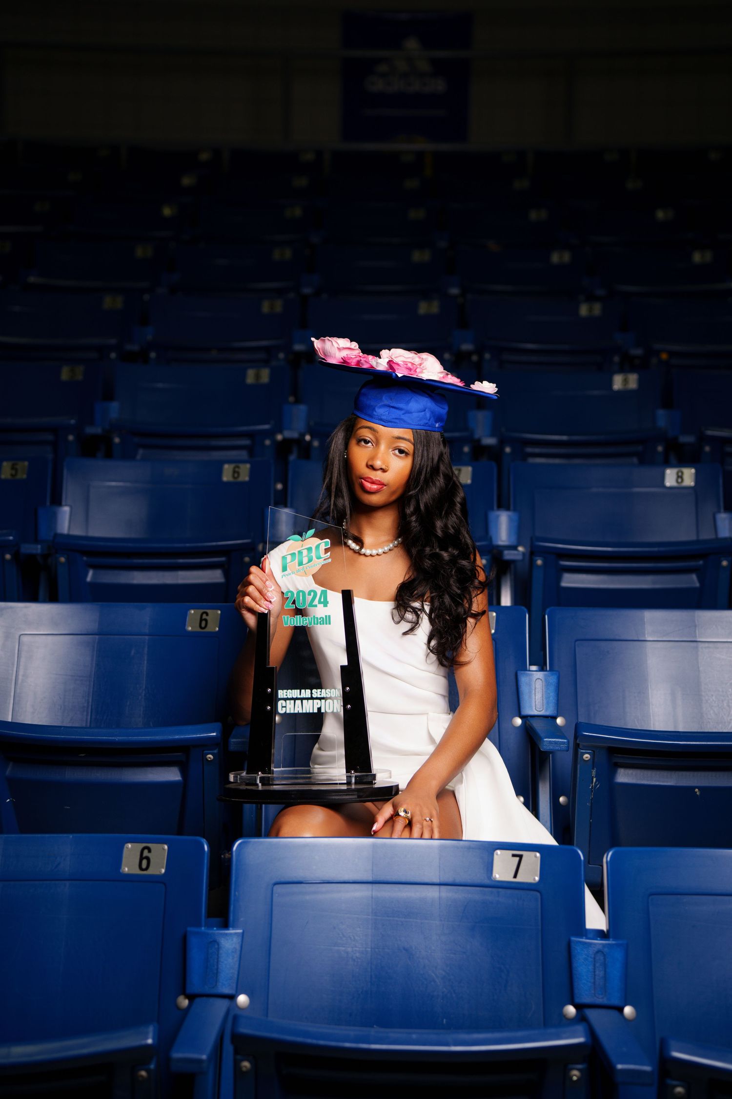 A young woman in a blue graduation cap and white dress holds a champion trophy while seated in stadium bleachers.