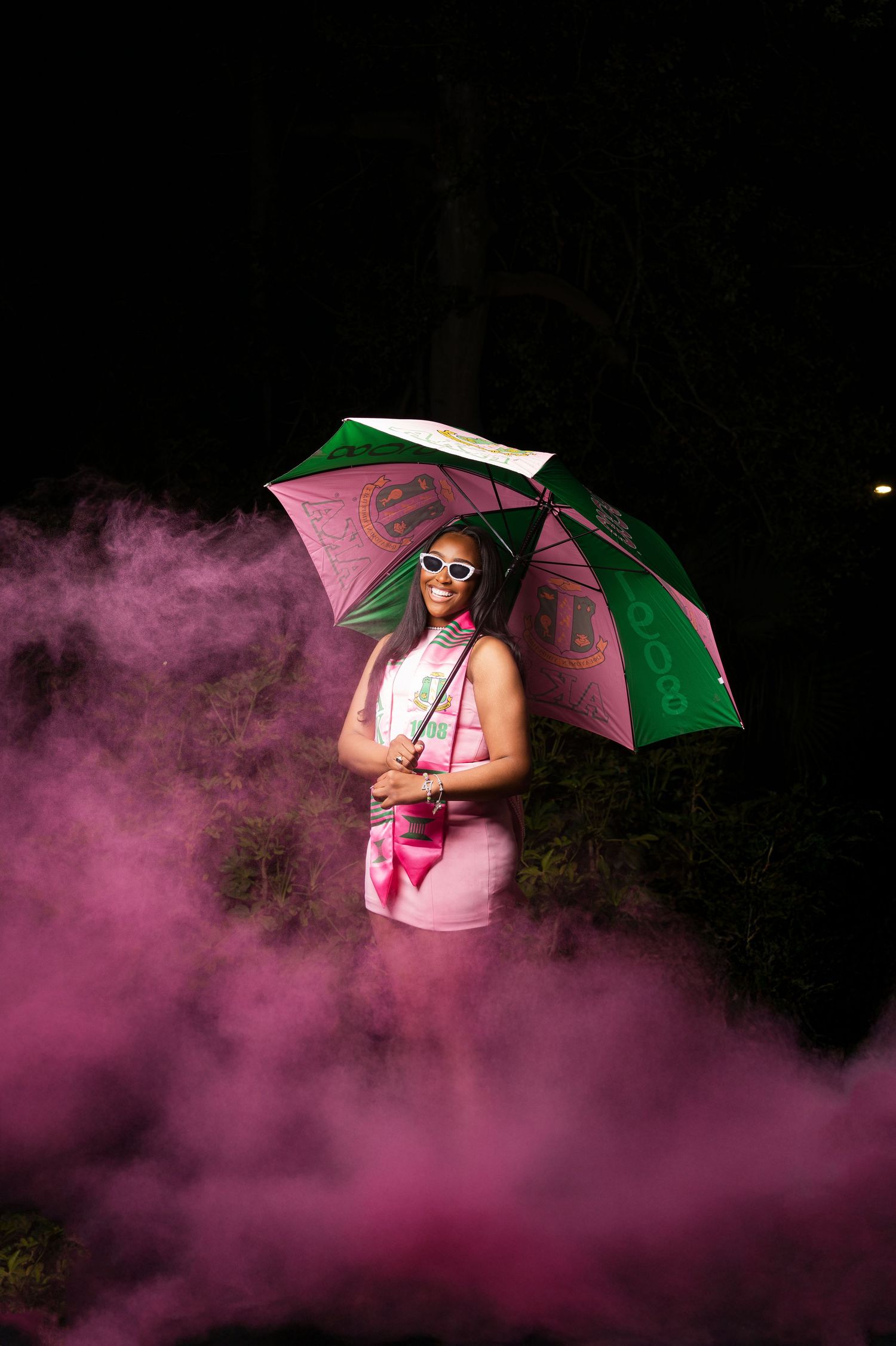 Woman in pink dress holding green and pink umbrella surrounded by pink smoke at night.