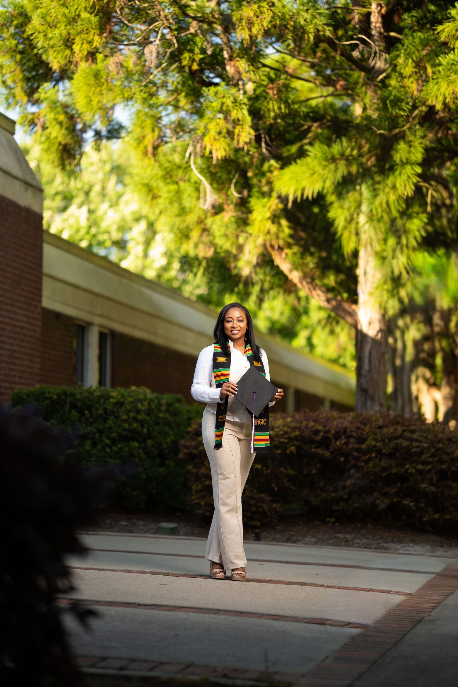 Young Black woman in graduation attire walks on campus path wearing kente stole and holding diploma.