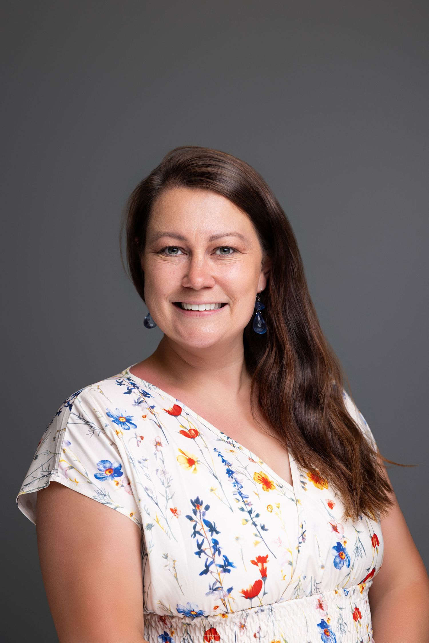 Smiling woman with long brown hair wearing a floral dress and blue earrings against a gray background.