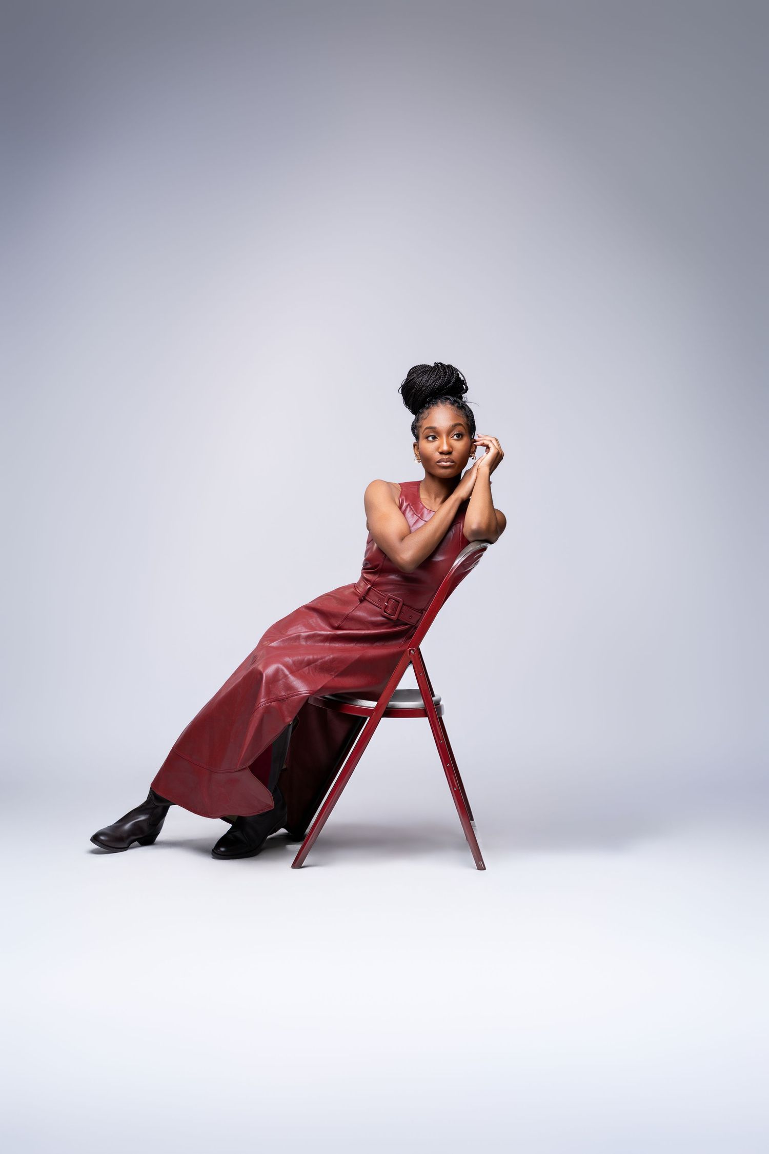 A figure in a burgundy dress poses dramatically on a red chair against a white studio backdrop.