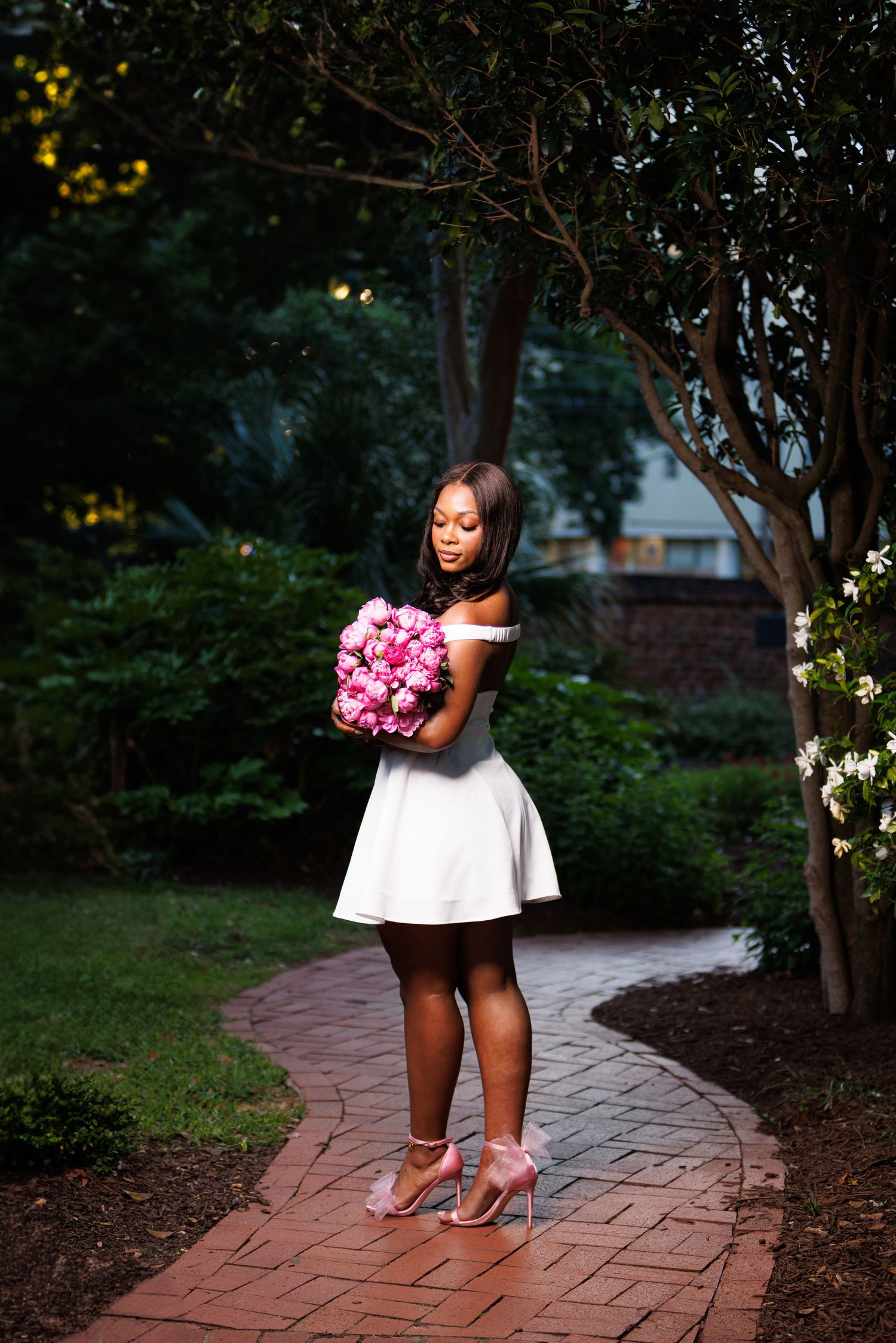 Woman in white off-shoulder dress holding pink flower bouquet on brick garden path.