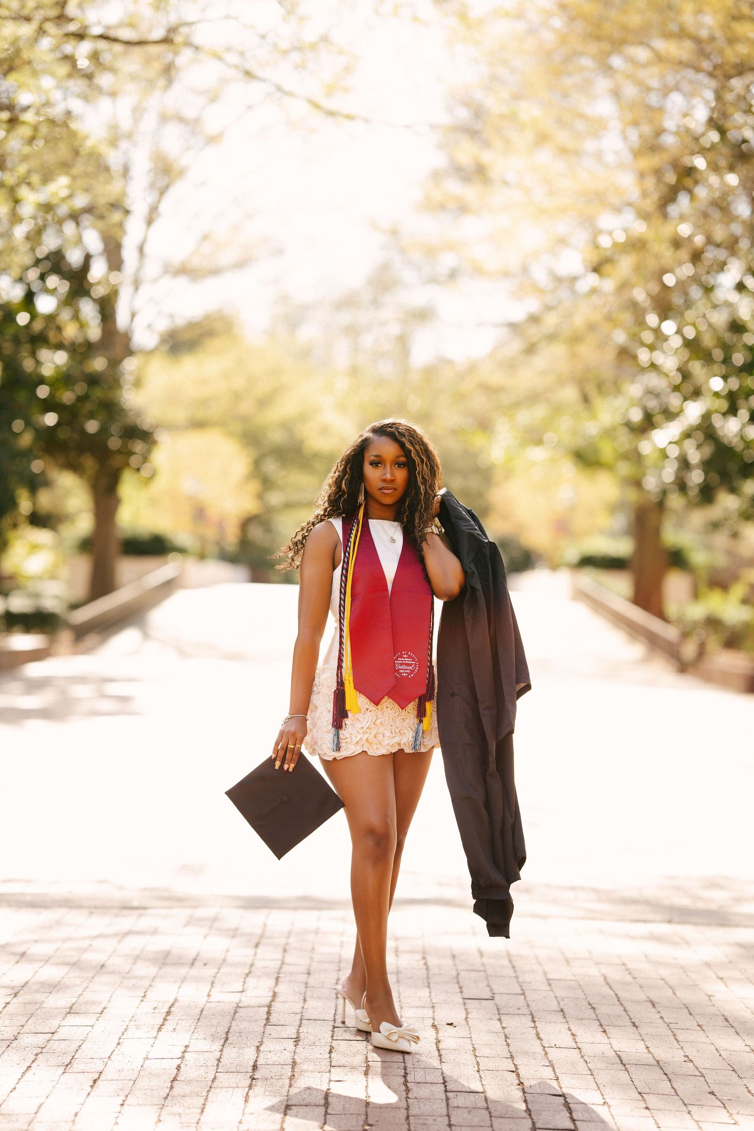 Young Black woman in graduation attire walks tree-lined brick path holding cap and gown on sunny day.