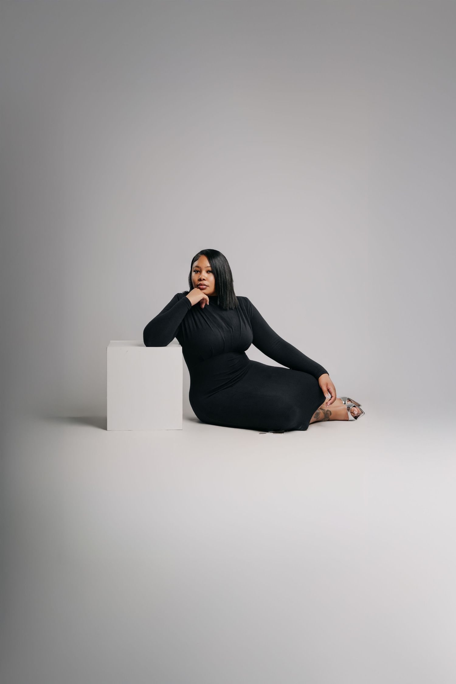 A plus-size woman in a black long-sleeve midi dress sits gracefully on a white cube against a gray studio background.