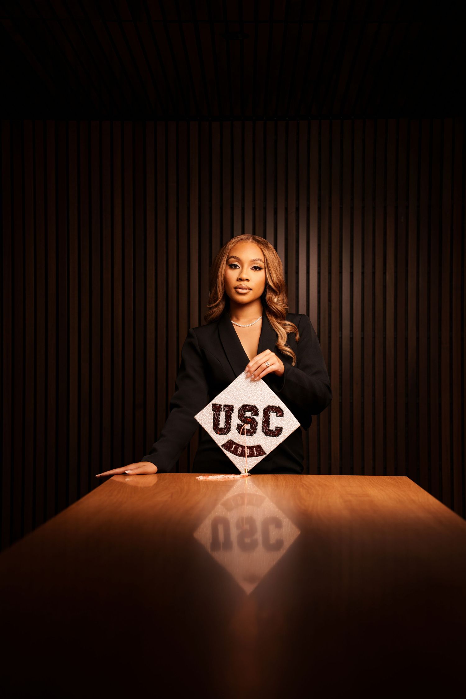 Woman in black blazer holding USC graduation cap at wooden table in dramatic dark studio setting.