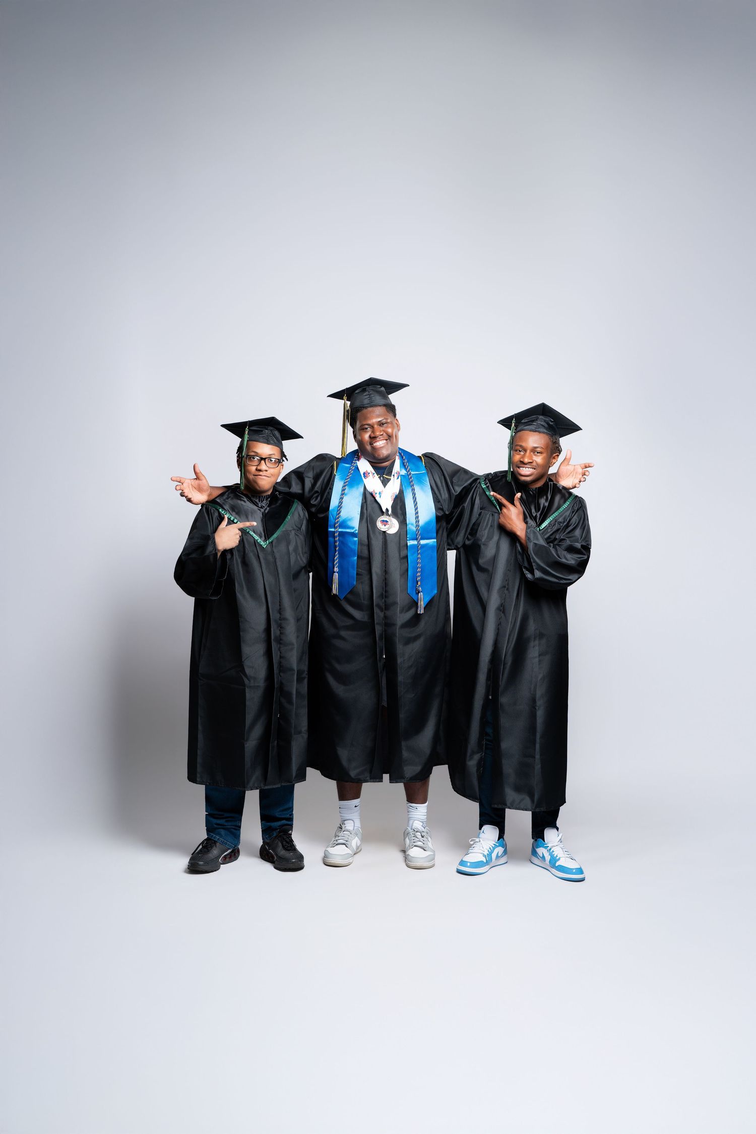 Three graduates in black caps and gowns pose proudly, with the center graduate wearing a blue sash and medal.