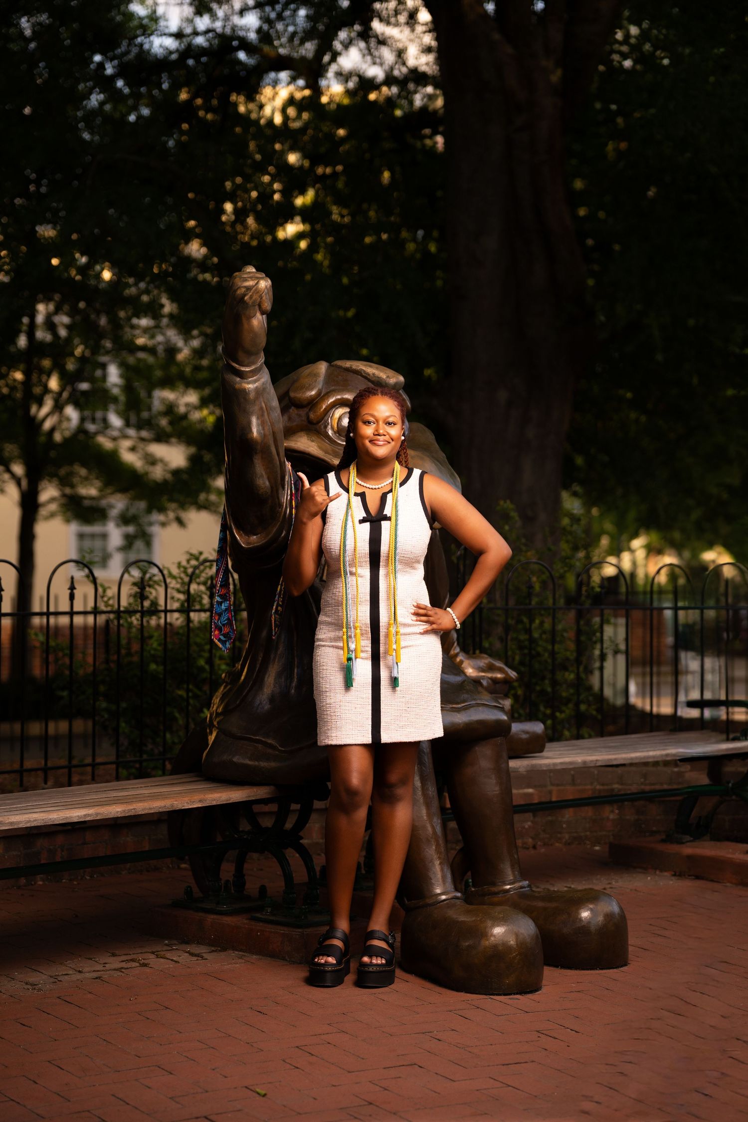 Young woman in white dress with colorful graduation regalia poses beside a bear statue on a brick plaza.