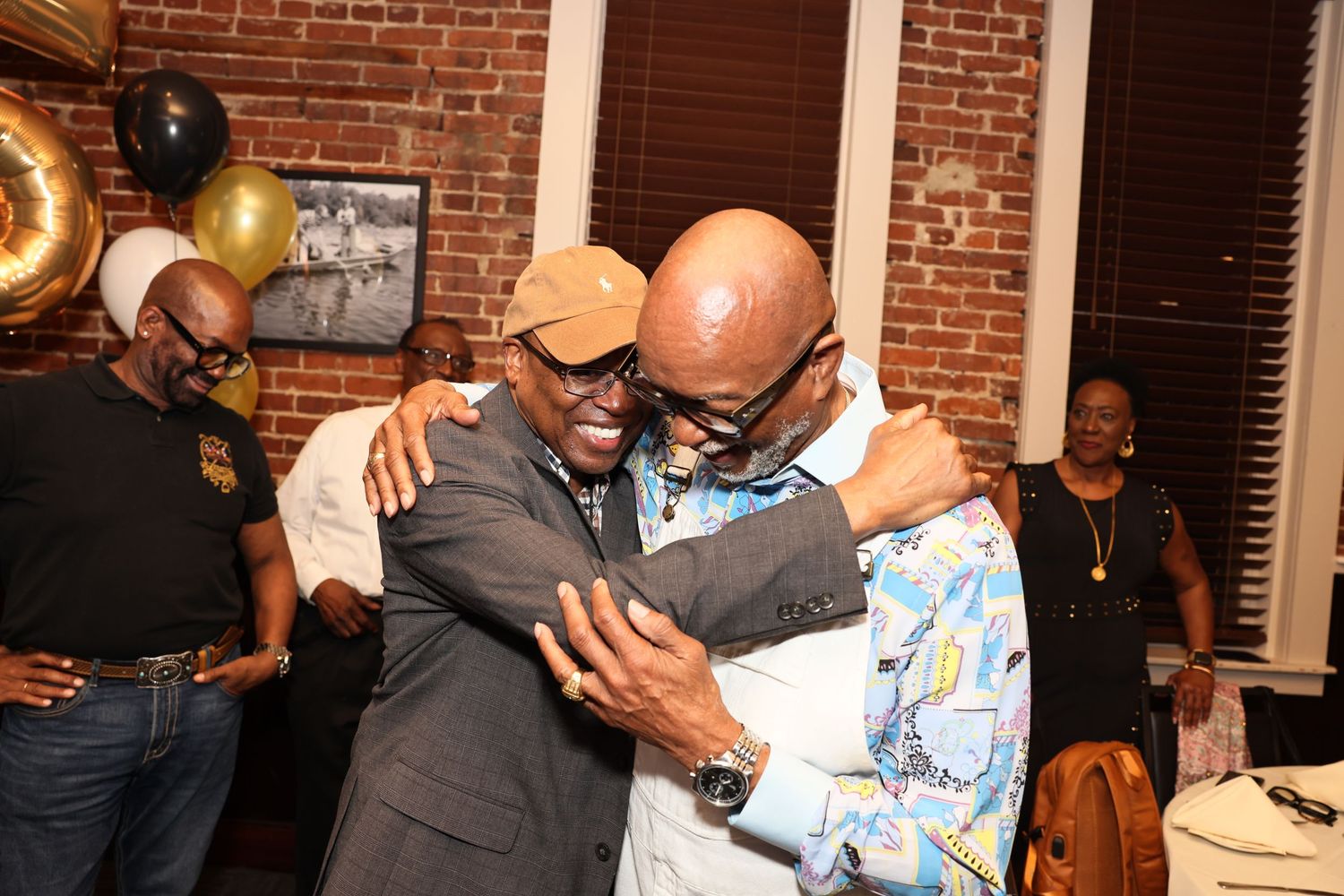 Two men share a warm hug at a festive party with gold and black balloons in the background.