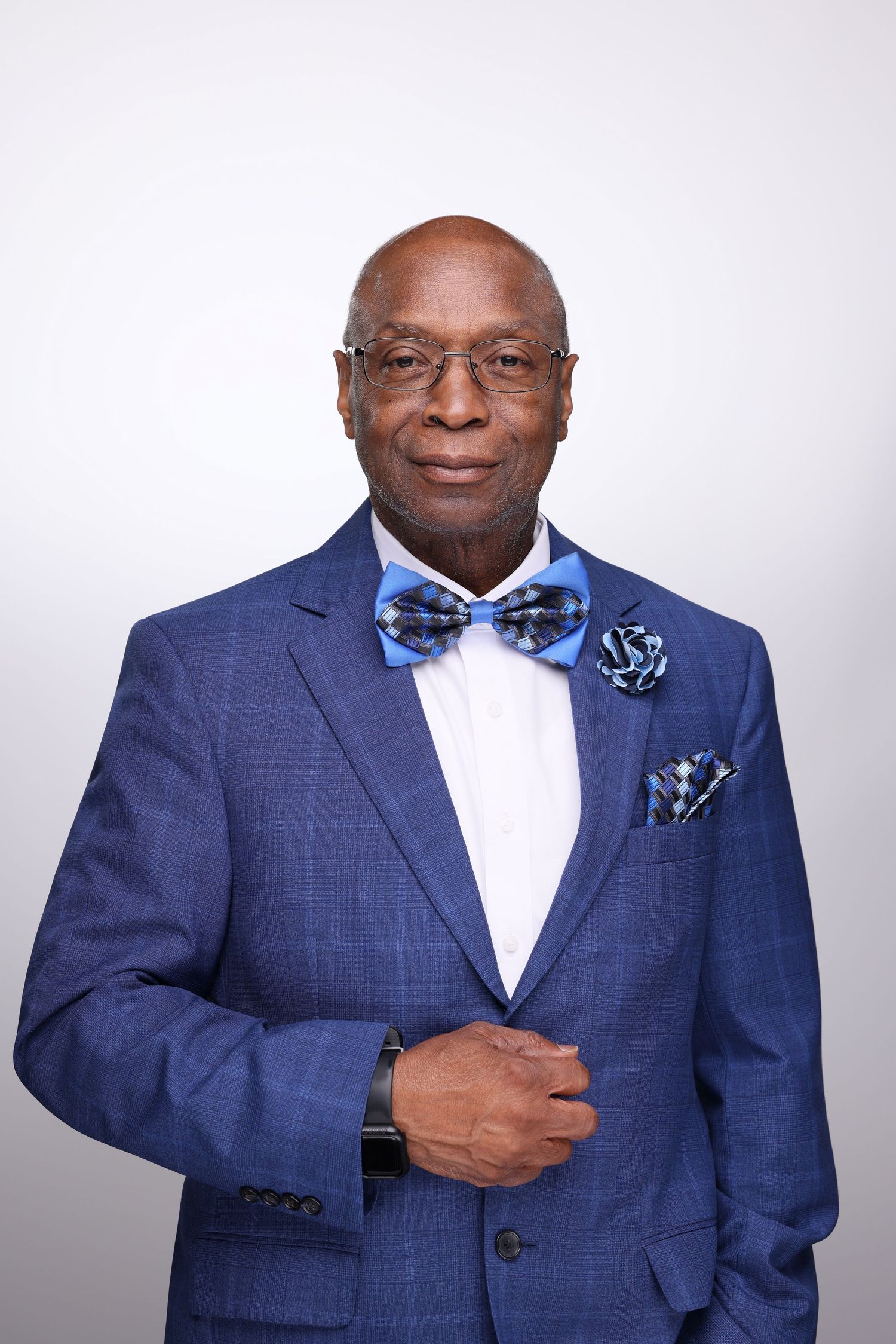 Elegant senior man in blue plaid suit, bow tie, pocket square, and glasses posing confidently against gray background.