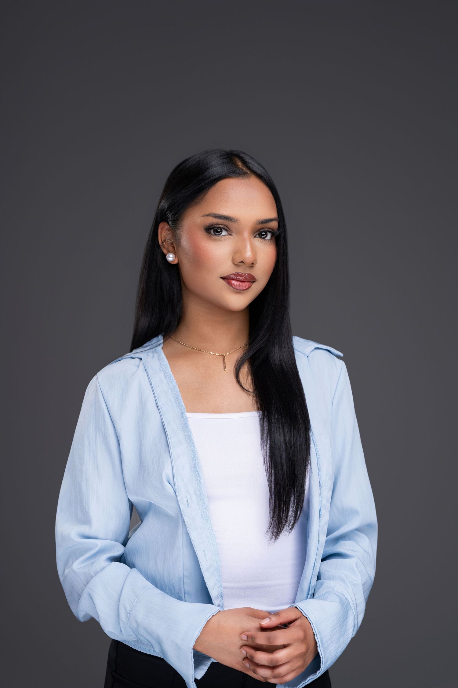 Young woman with long black hair wearing a light blue shirt over white top, posing confidently against gray background.