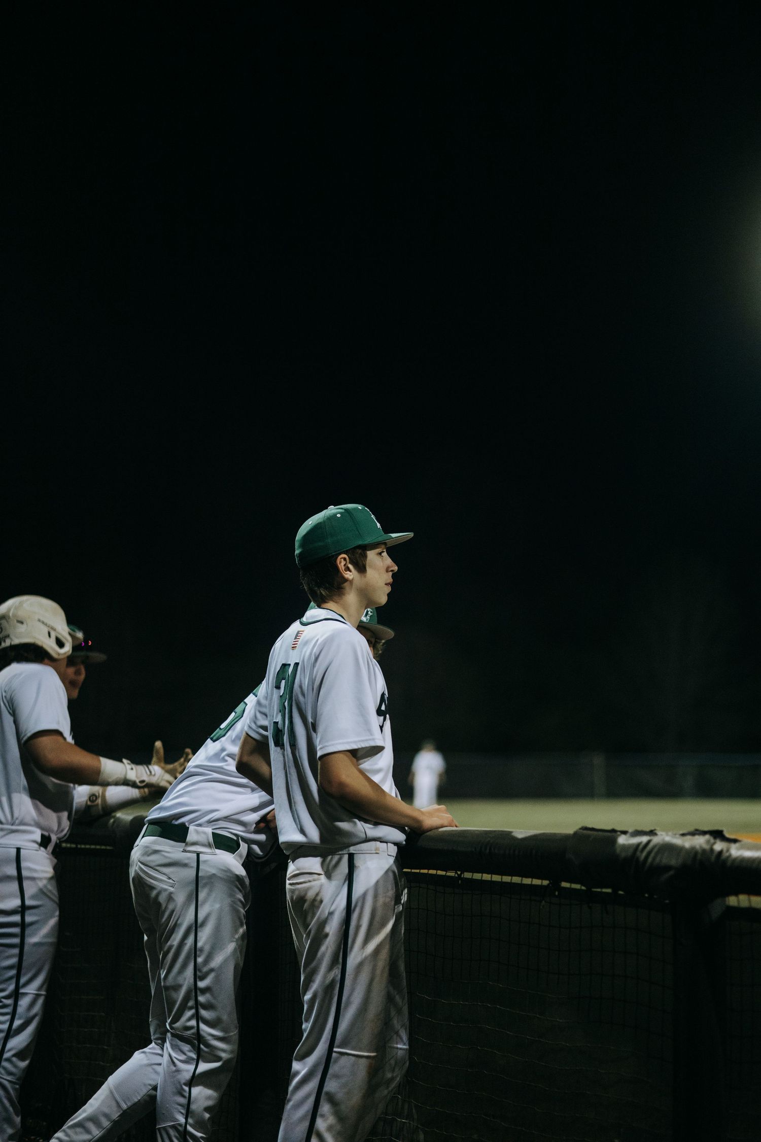 Baseball players in white uniforms lean against dugout railing during nighttime game.