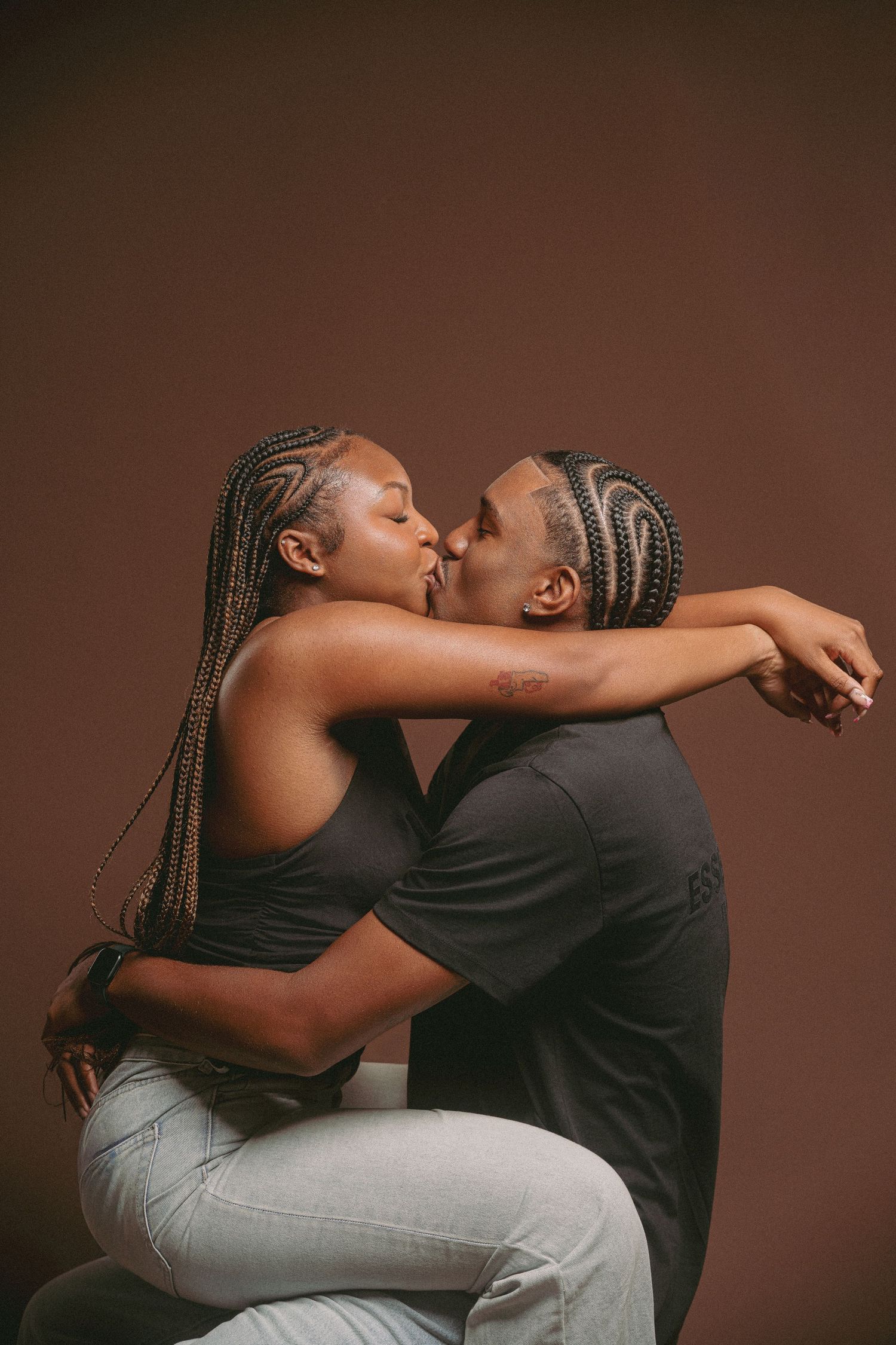 A couple sharing a romantic kiss in a studio portrait, both wearing casual outfits with braided hairstyles.