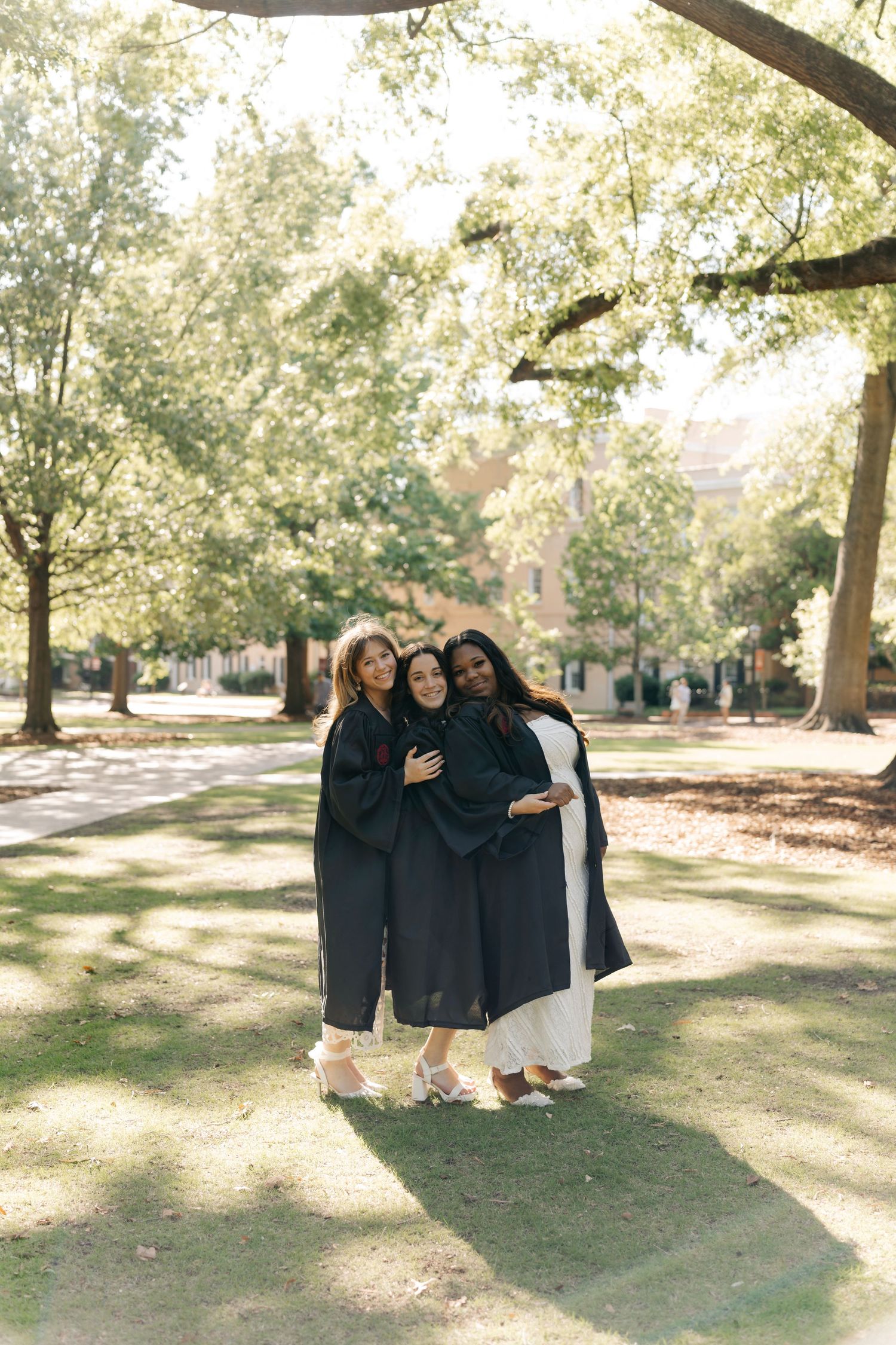 Three graduates in black gowns pose together outdoors on a sunny campus lawn surrounded by green trees.