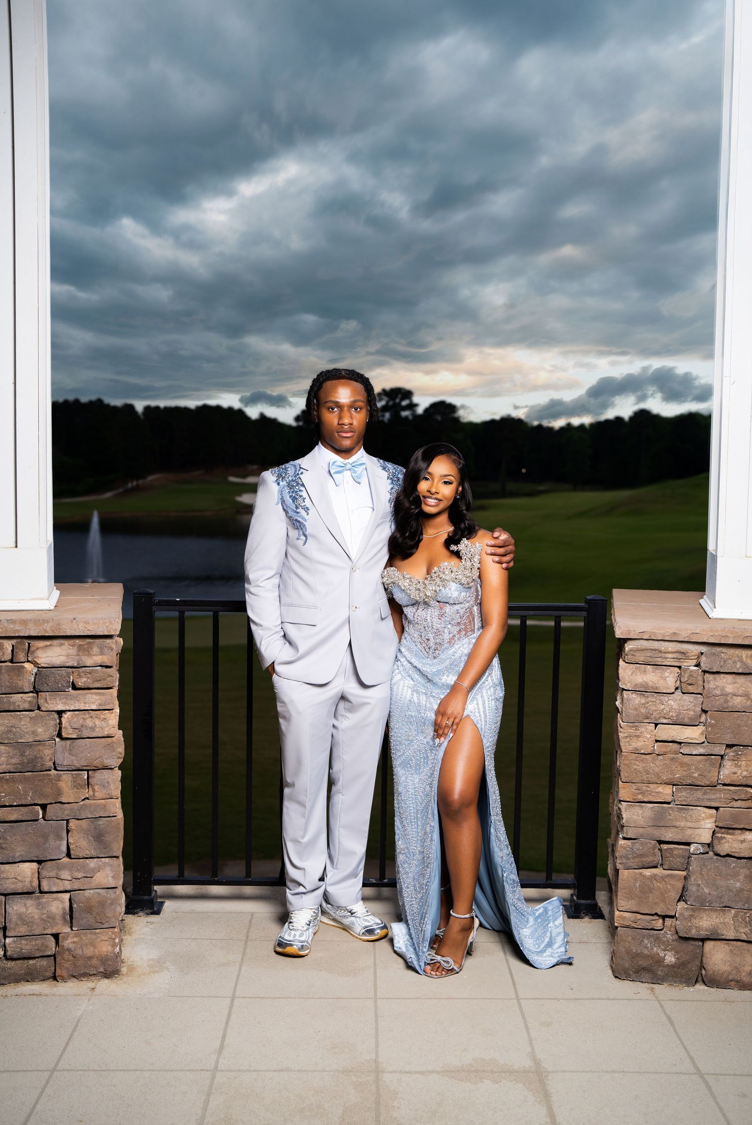 A young couple poses in coordinating light blue prom attire on a stone terrace overlooking a golf course at dusk.