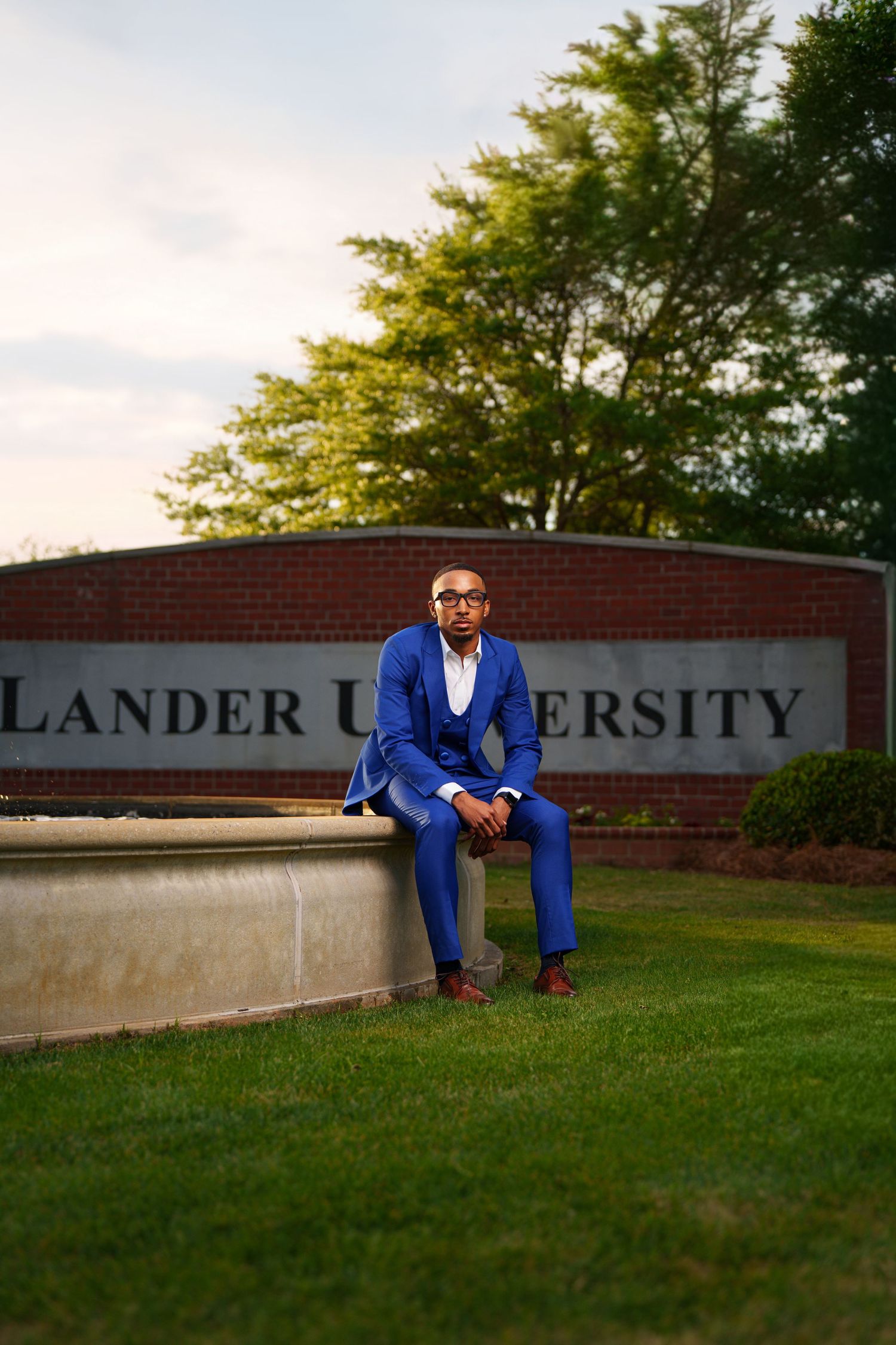 A man in a royal blue suit sits confidently in front of Lander University's brick entrance sign on a green lawn.