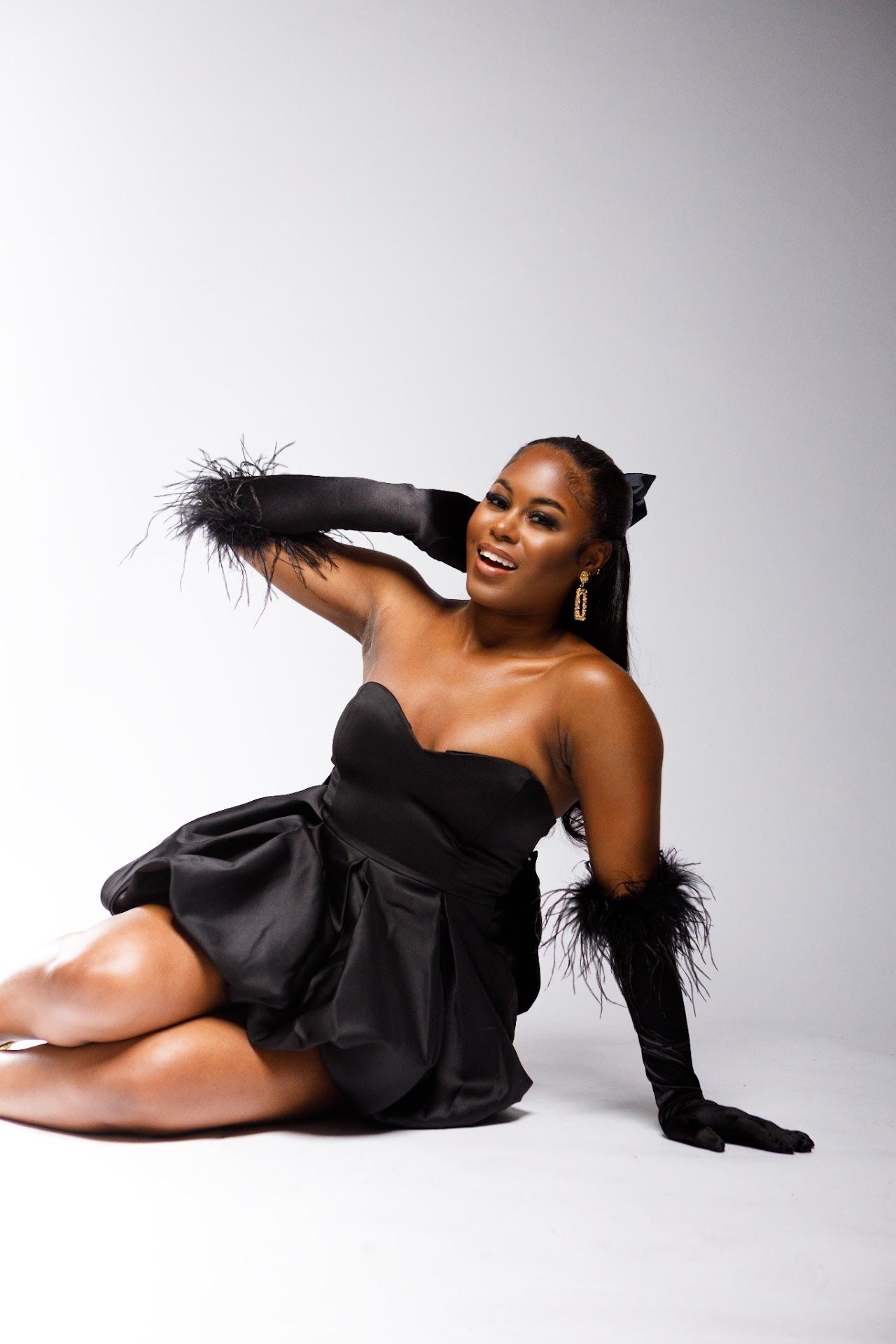 Fashion photo of a model in an elegant black strapless dress with feather details posing on a light gray background.