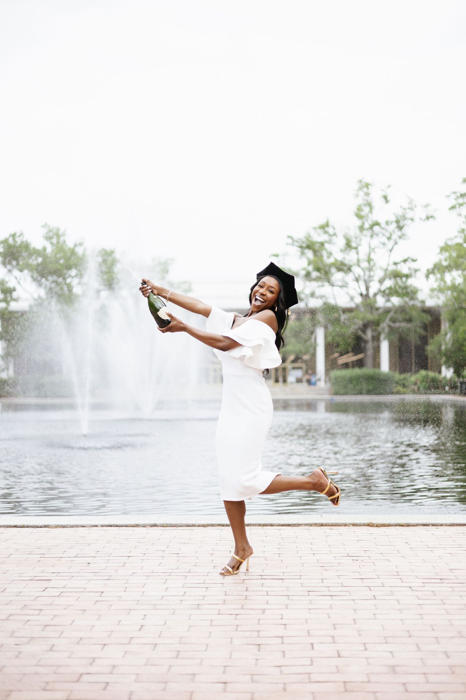 Graduation photos showing joyful poses in a white dress by a fountain on a brick path.