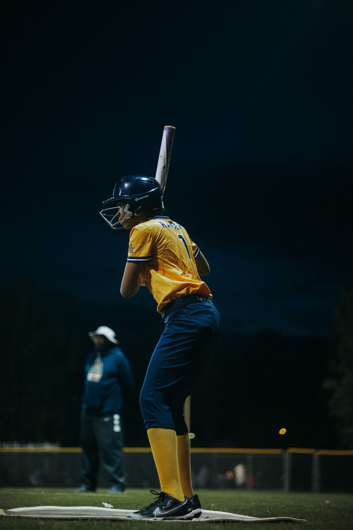 Softball batter in yellow uniform takes stance at plate during evening game.