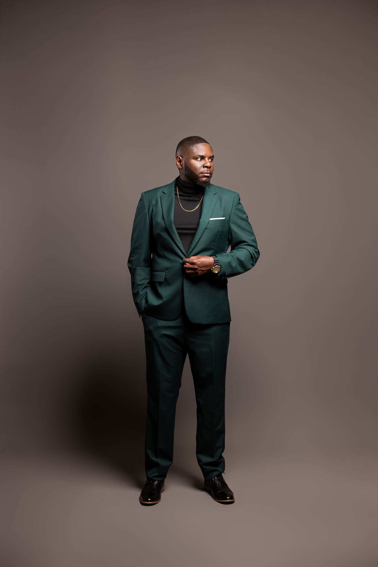 Professional portrait of a person in an emerald green suit and black turtleneck standing against a gray backdrop.