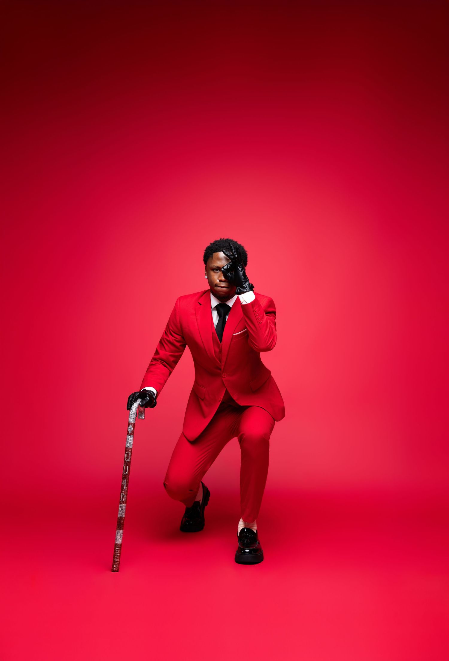 Man in red suit holding a cane against a red background, striking a dramatic pose.