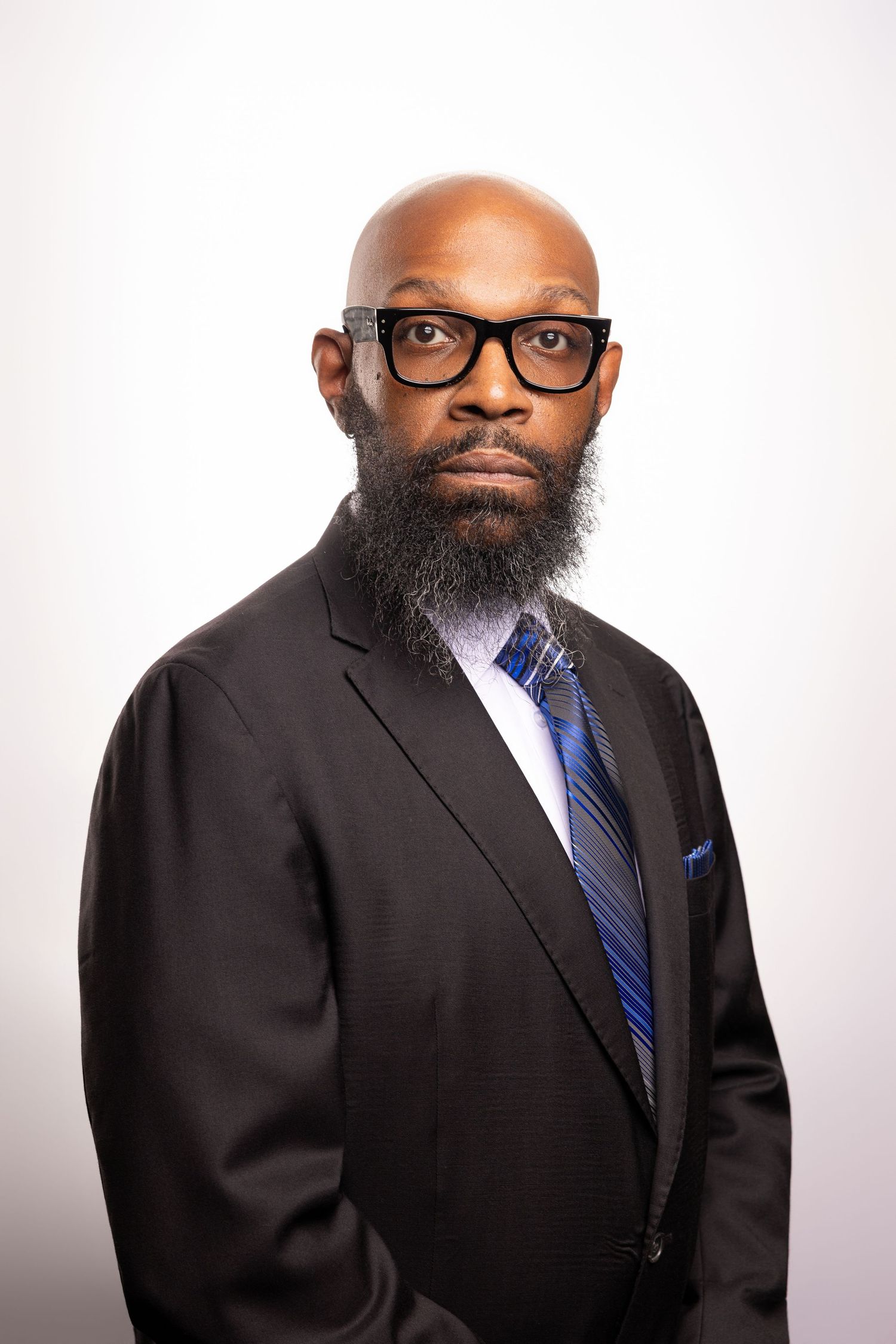 Professional bald Black man with beard wearing black suit, blue striped tie, and glasses against white background.