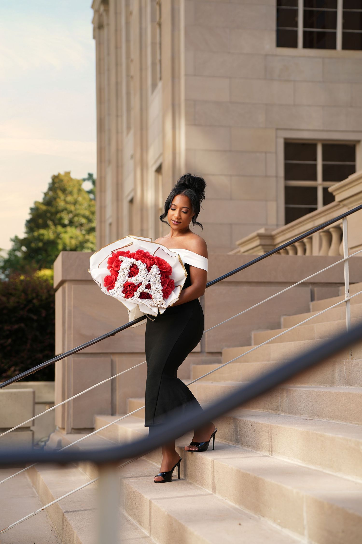 Elegant woman in black off-shoulder gown holding red rose bouquet spelling PA on stone steps.