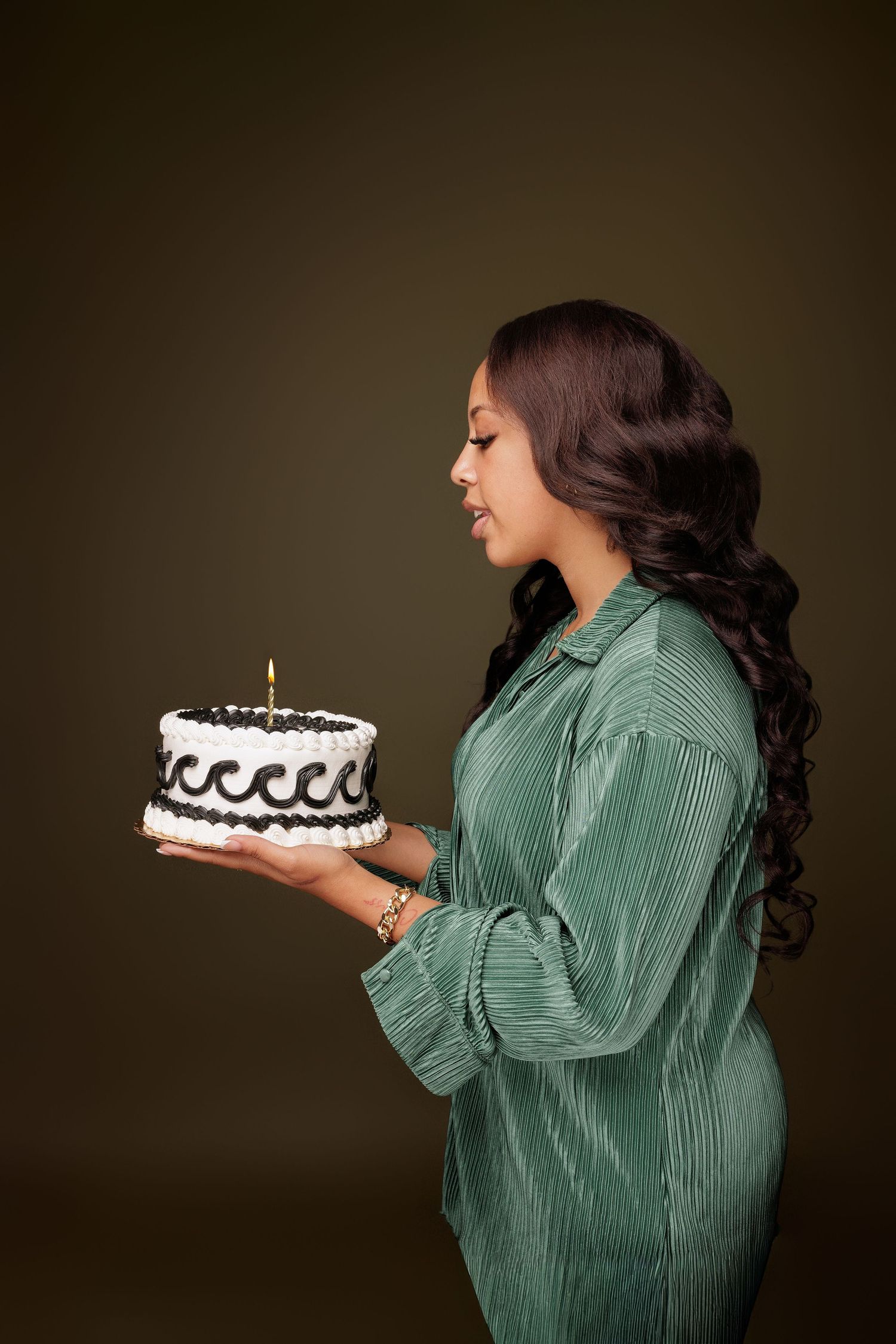 A person in a green corduroy shirt holds a black and white birthday cake with a lit candle against a dark background.