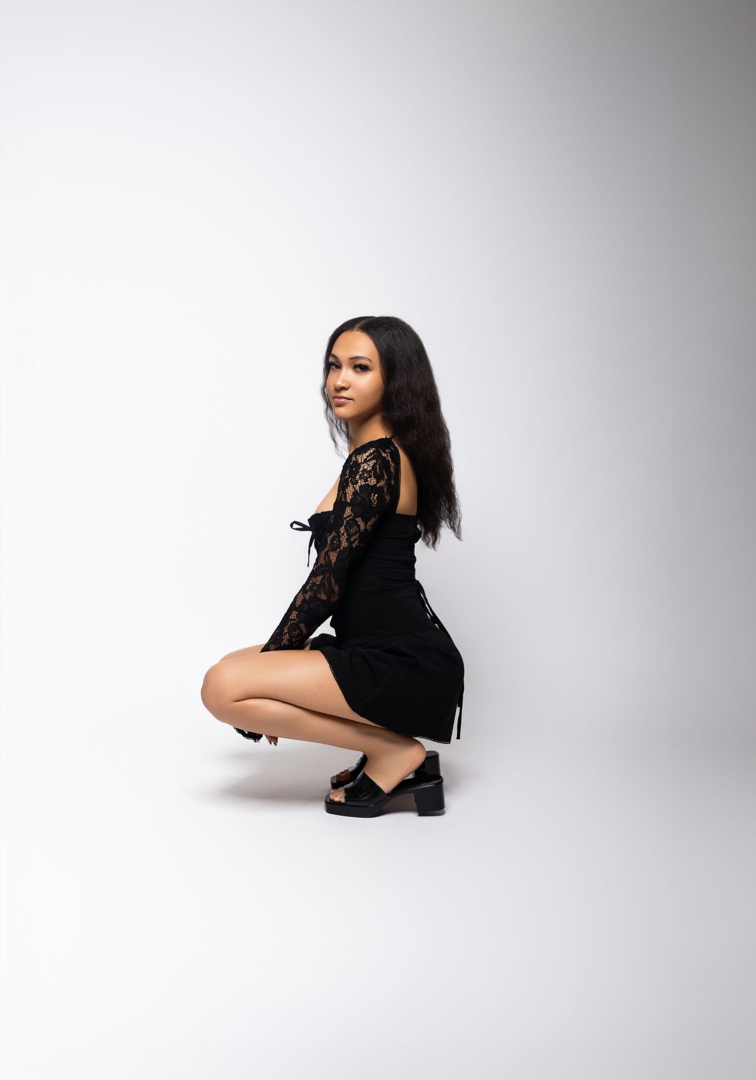 Fashion portrait of model in black lace dress crouching against white background.