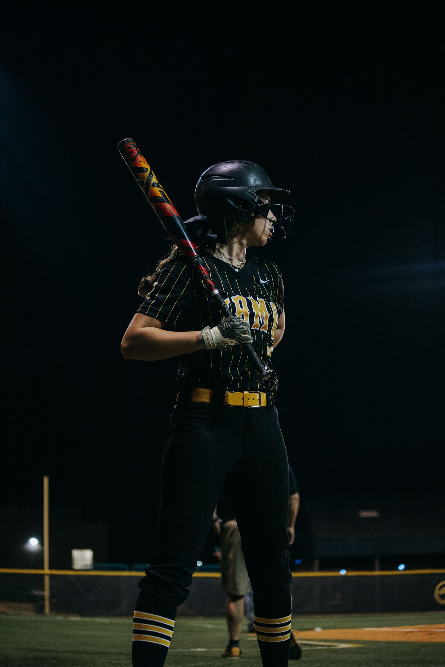 Softball player in black and gold uniform holds bat at ready position.