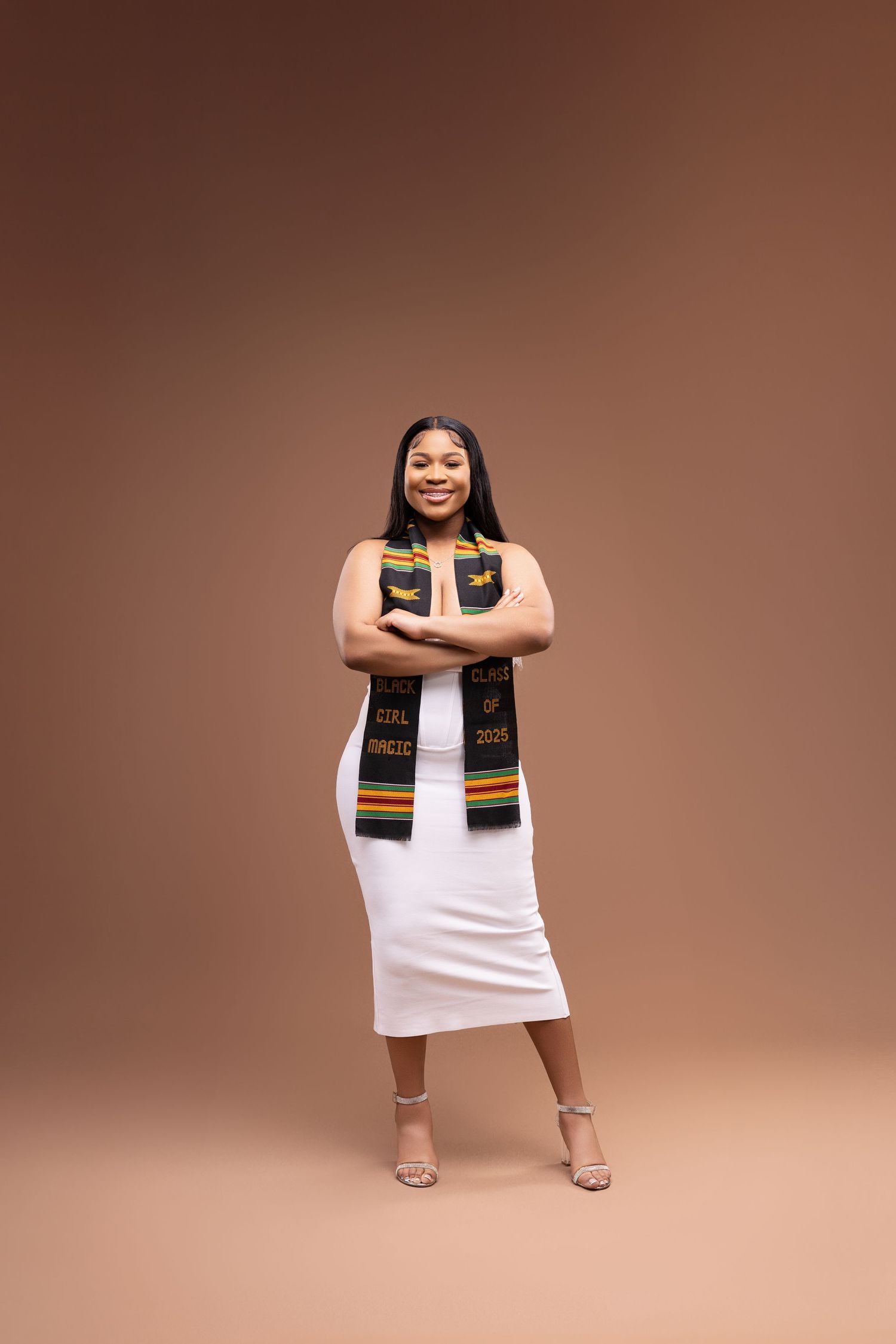 Graduate wearing white dress and colorful graduation stole poses confidently against brown background.